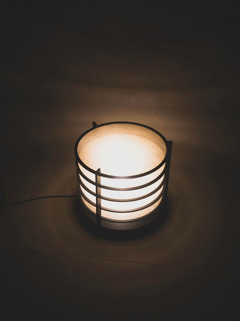 The Sculptural Lamp by ELIURPI features a fabric shade and horizontal wooden slats, emitting ambient light in a dark room to create a cozy atmosphere. Its cord is visible on the left side, adding to its sculptural appeal.