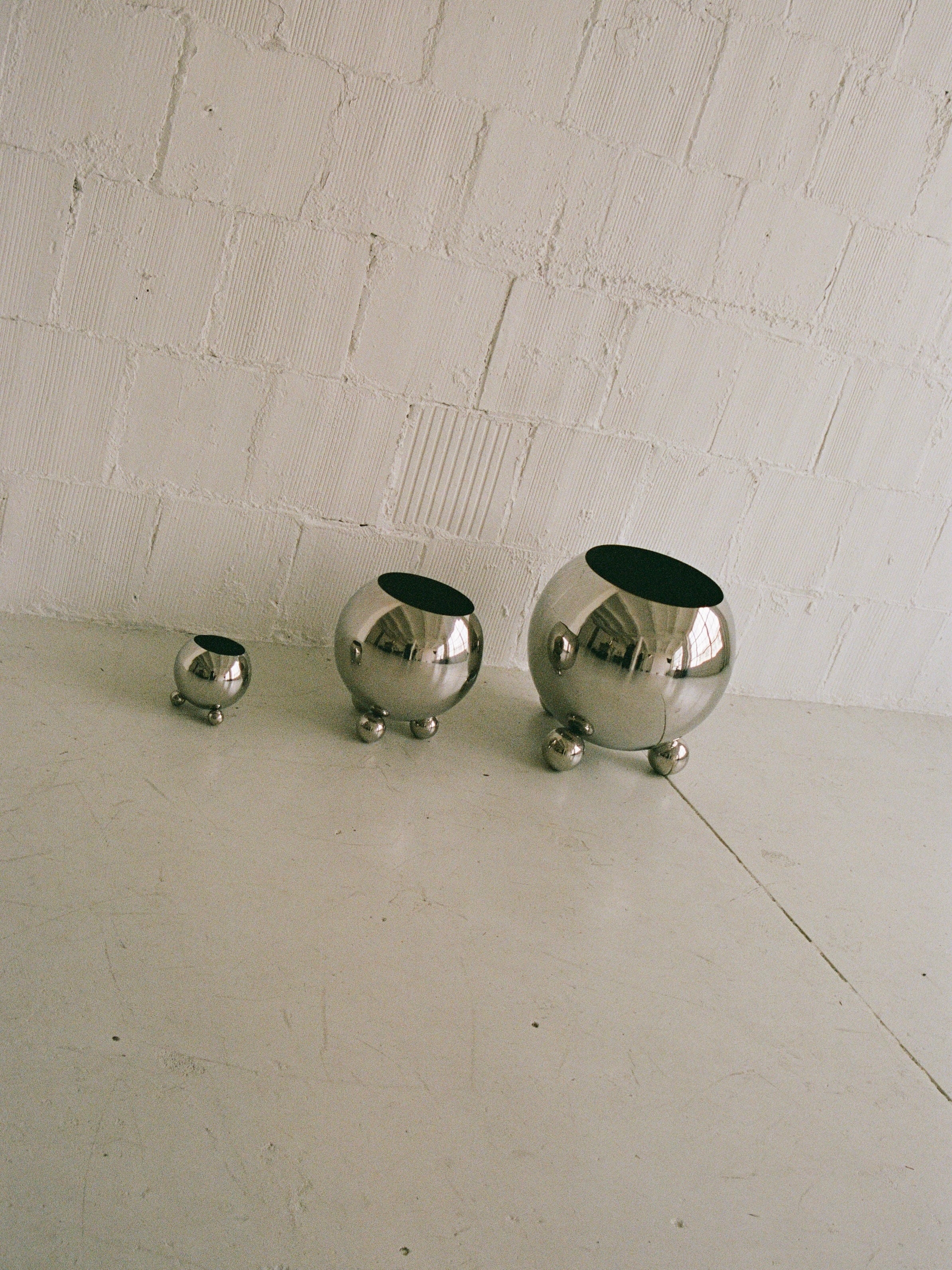 Three reflective Selene Planters from MUHLY, made of stainless steel and available in varying sizes, are aligned on a light-colored floor against a white brick wall. These spherical planters feature round, ball-like feet that contribute to their sleek, modern look. Arranged from smallest on the left to largest on the right, they create a striking display.