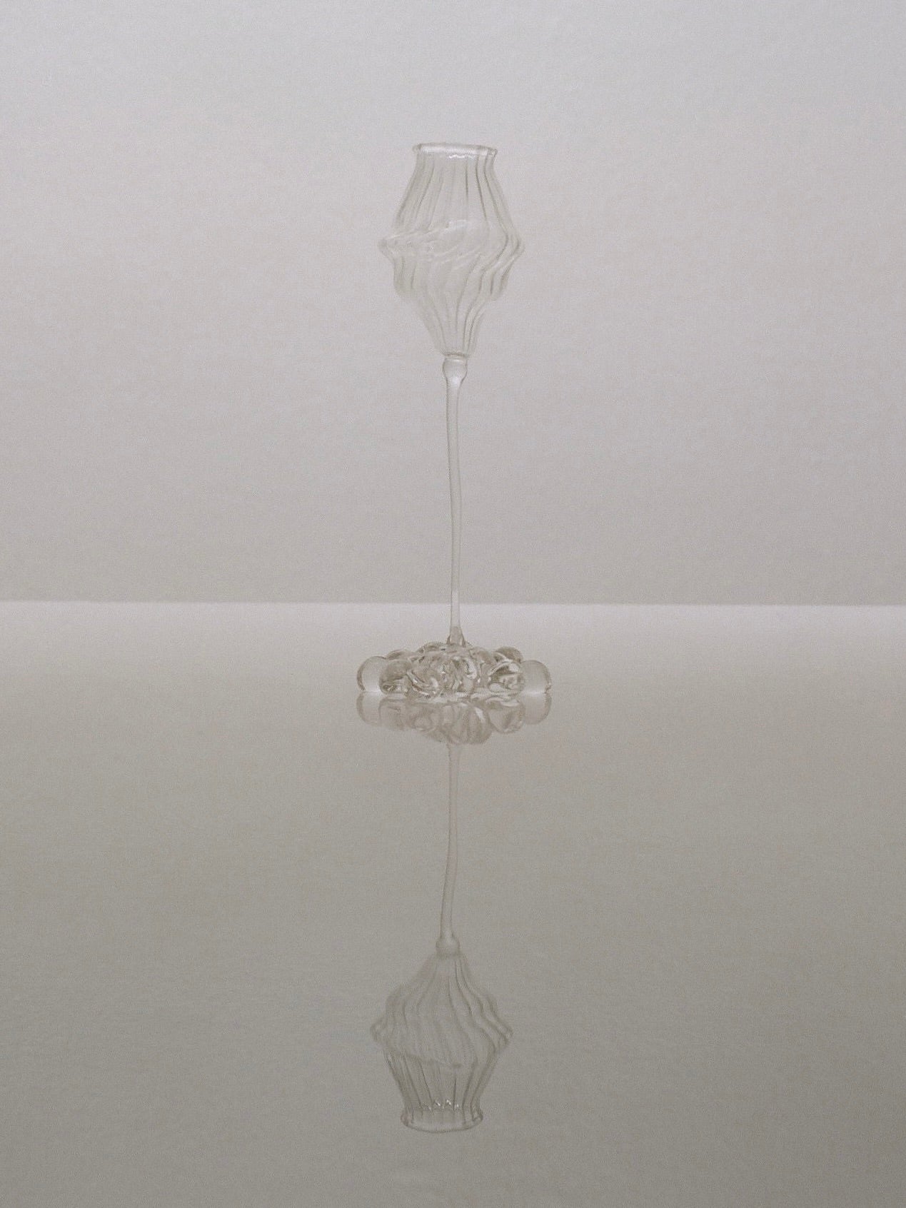 Standing on a reflective surface is an intricately designed, tall, transparent glass sculpture with a wavy, textured surface. This elegant piece, with its mouth-blown form and perfect symmetry, creates a tranquil and balanced visual effect reminiscent of the Orphée Liqueur Glasses by Justine Menard.