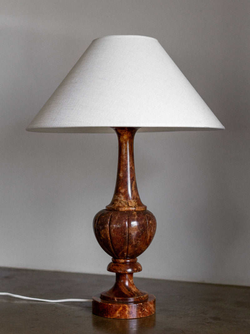 A Large Italian Alabaster Stone Lamp by Spigel, featuring a wooden base with a hand-carved design and topped with a white fabric lampshade. The lamp, exuding neoclassical style, is placed on a surface against a plain wall, with its white electrical cord visible at the base.