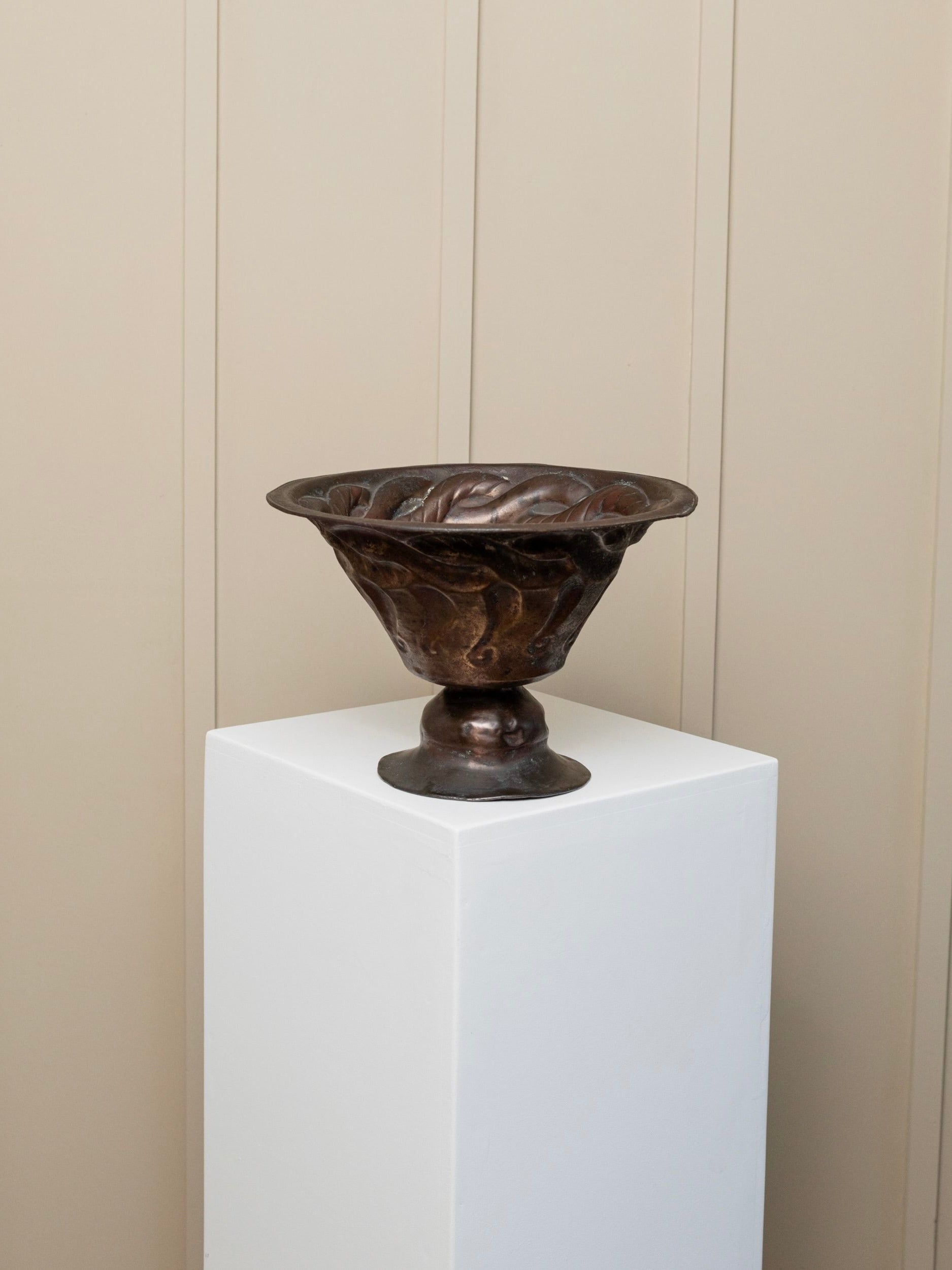 An Antique Fruit Bowl - Hand-Carved Copper by Bottega Jacobs is displayed on a white pedestal against a plain beige wall, highlighting its exquisite antique patina.