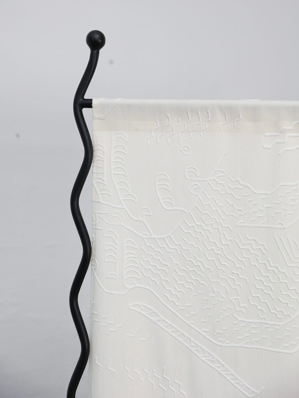 Close-up of The Good Living&Co.'s "Alto" Folding-Screen, featuring handcrafted white fabric with a raised abstract pattern hanging from a wavy black metal rod, strikingly prominent against a plain light gray background.