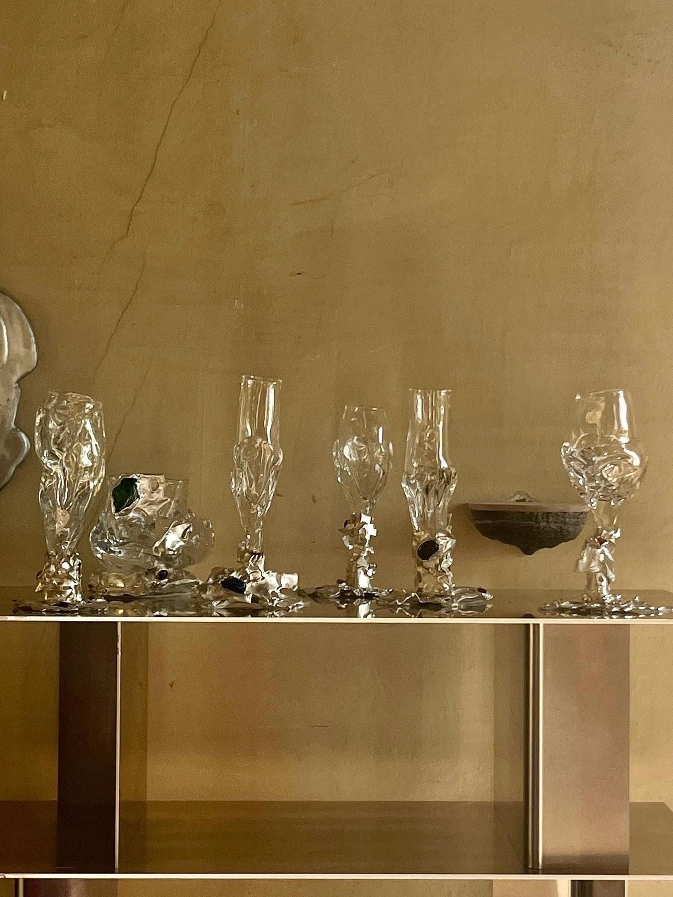 A display shelf showcases a collection of meticulously crafted glassware, featuring hand-blown pieces such as the Jewelled Goblet no. 10 by Lindsey Fontijn from Szkło Studio. Each piece boasts an individual artistic design with twisted stems and intricate bases. The background is a simple, light brown wall that has some noticeable cracks.