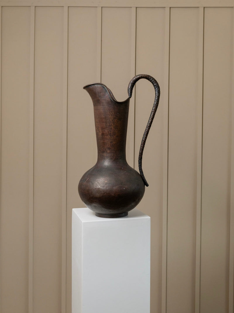 A tall vintage metal jug by Bottega Jacobs is elegantly placed on a white pedestal against a beige paneled wall, evoking the artistry of handcrafted Italy from the 1950s.