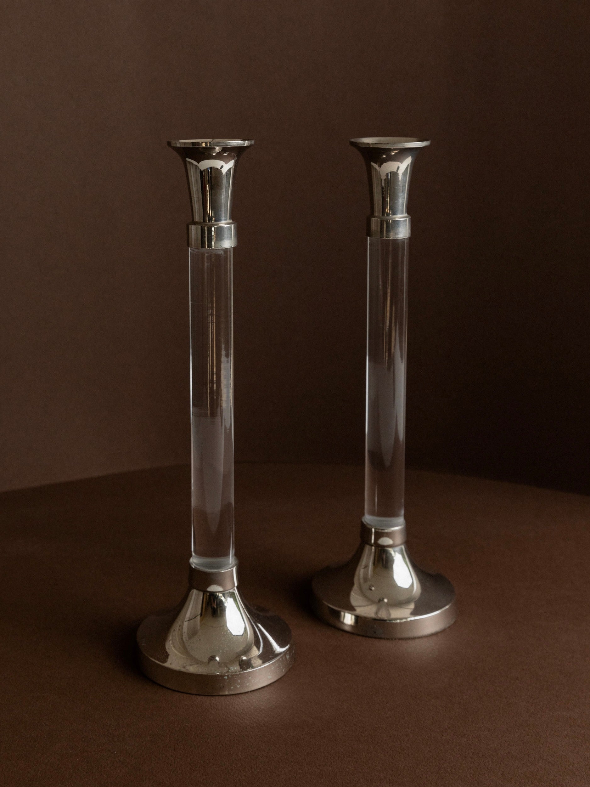 The Set of 2 Candleholders by Bottega Jacobs are positioned on a brown surface against a brown backdrop. These candleholders exhibit a sleek and modern design, showcasing chrome-plated metal bases and tops connected by an acrylic glass column.