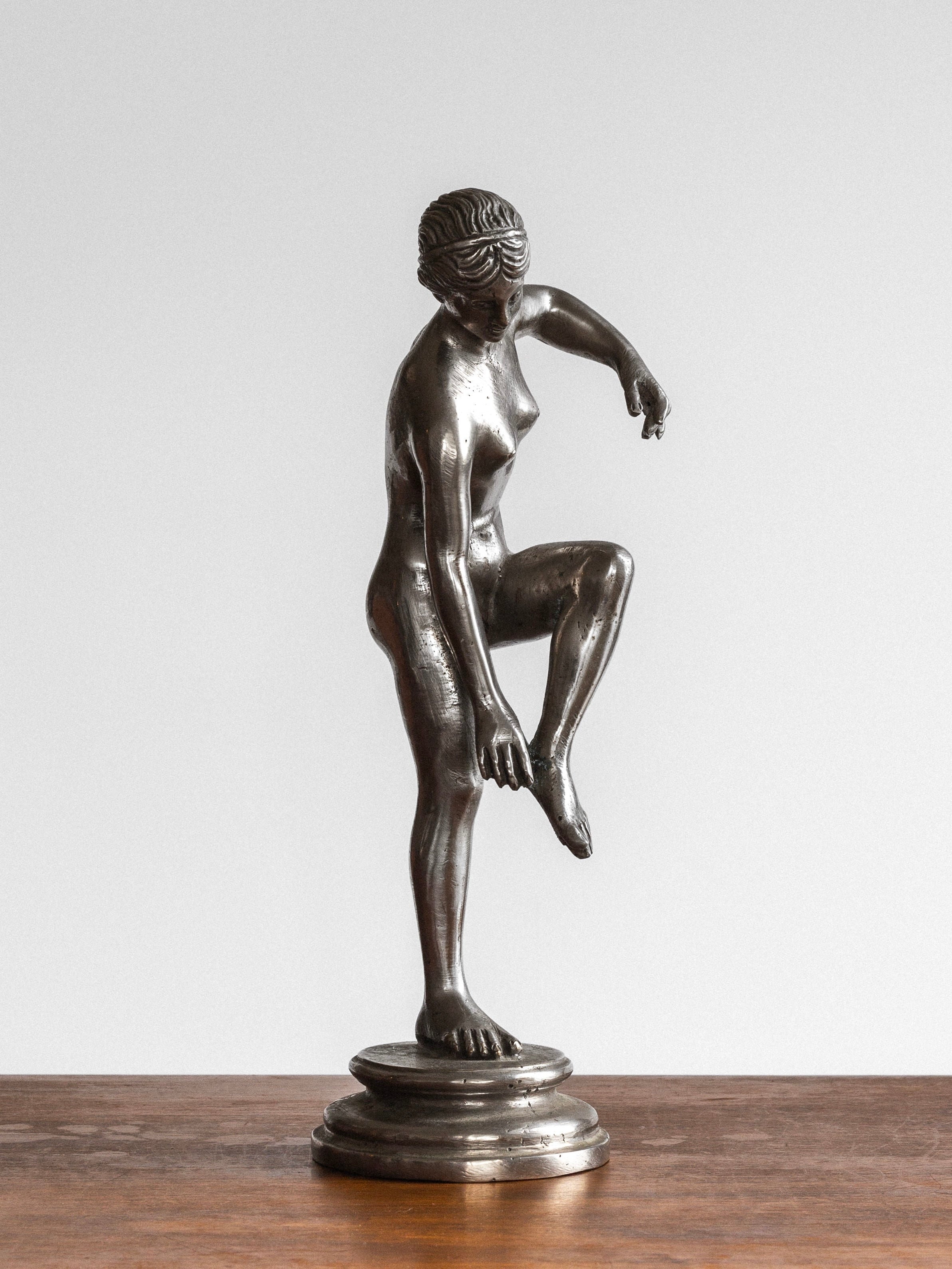 The "Elegant Metal Sculpture, 1930s" by Spigel captures a neoclassical design with a bronze statue of a woman featuring intricately detailed hair. She stands on one leg while raising the other and points downward with both hands toward her raised foot. The statue is mounted on a round base and elegantly displayed on a wooden surface.