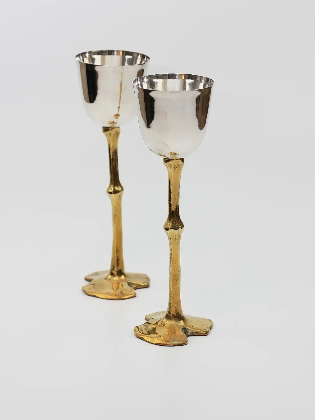 A pair of Brutalist Goblets from Boga Avante Shop, featuring elegant silver-colored cups and tall, slender brass stems and bases, is showcased against a plain white background. The smooth, reflective surface of the goblets contrasts beautifully with the textured, ornate stems that are hand-sculpted in a striking Brutalist style.