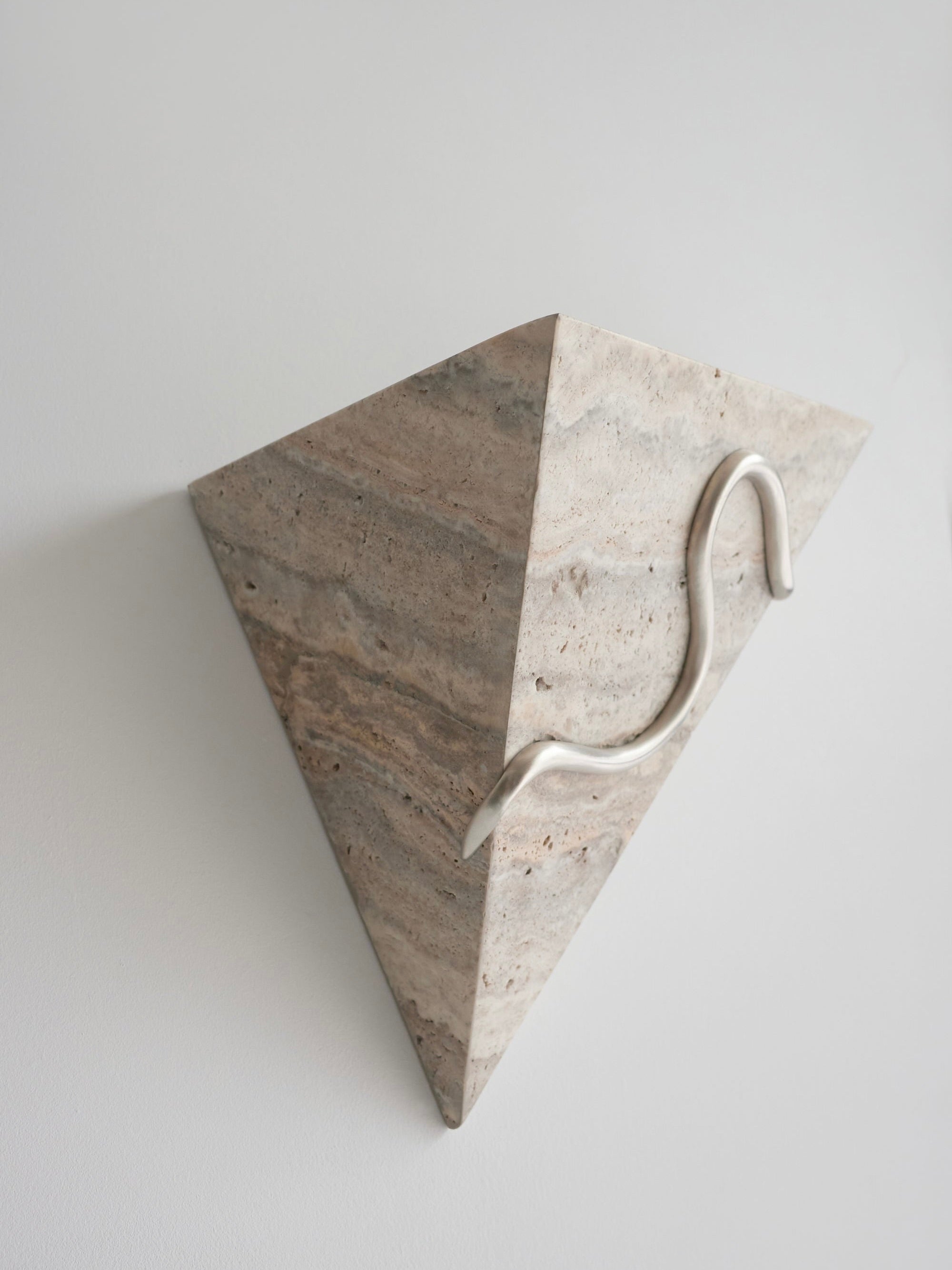 Introducing the Therapeutic Wall Light 3 by Mariza Galani: This sculptural wall piece showcases a triangular, stone-like form with a striated texture, elegantly mounted on a white wall. A handcrafted silver travertine element loops gracefully from the top to the right side, adding a modern and artistic touch.