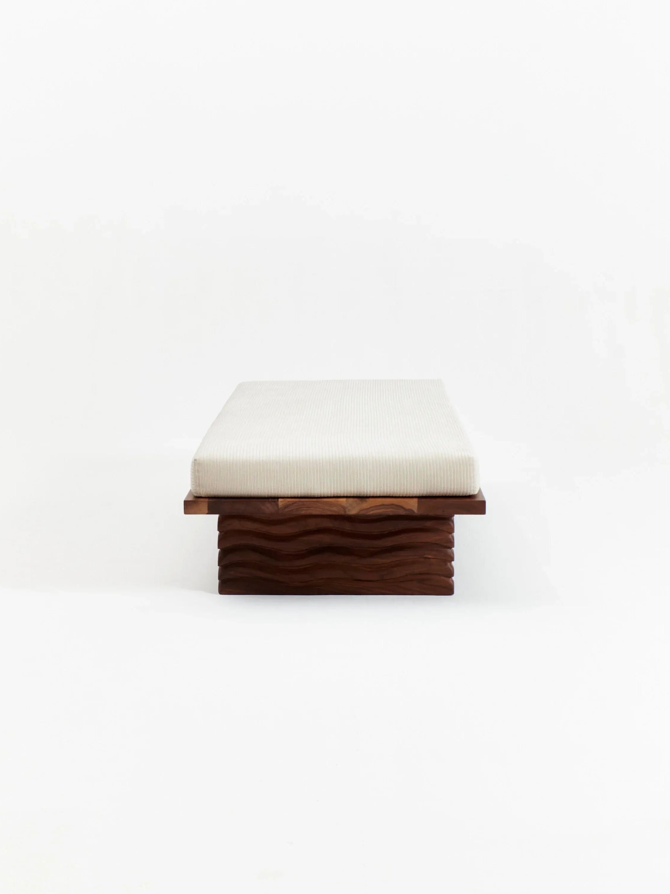 The Project 213A Nazaré Daybed is a minimalist wooden bench with a white cushioned seat, featuring a textured solid walnut base adorned with wave-like carvings for an artistic touch. Handmade in Portugal, the plain white background accentuates its clean lines and exquisite craftsmanship.