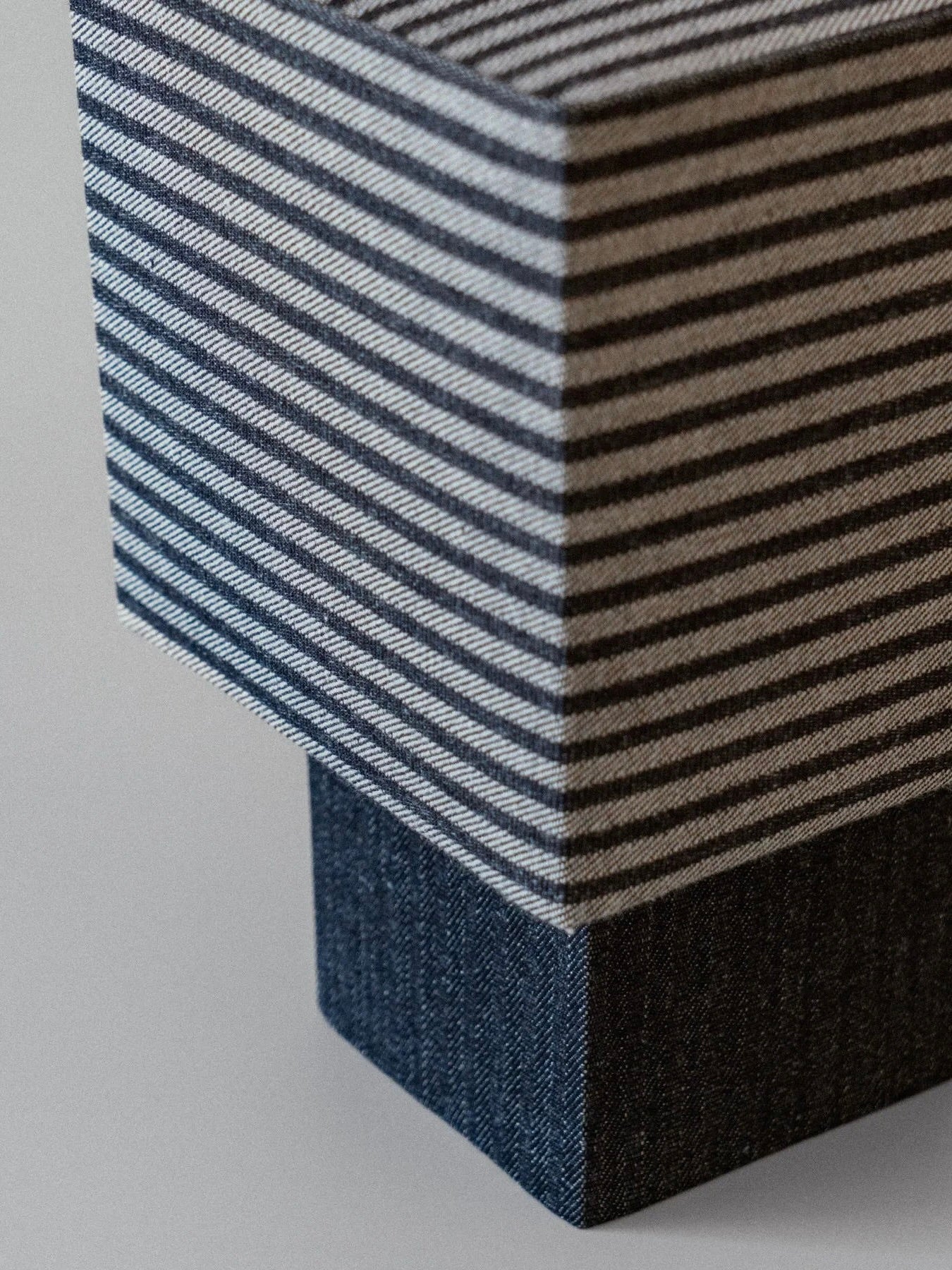 The "La Petite Boîte" lamp by mattina moderna features a close-up of its striped denim textile with alternating dark and light gray lines draped elegantly over the handcrafted lamp. The lower section is crafted from solid dark fabric using deadstock materials, creating a striking visual contrast with the sustainable pattern.