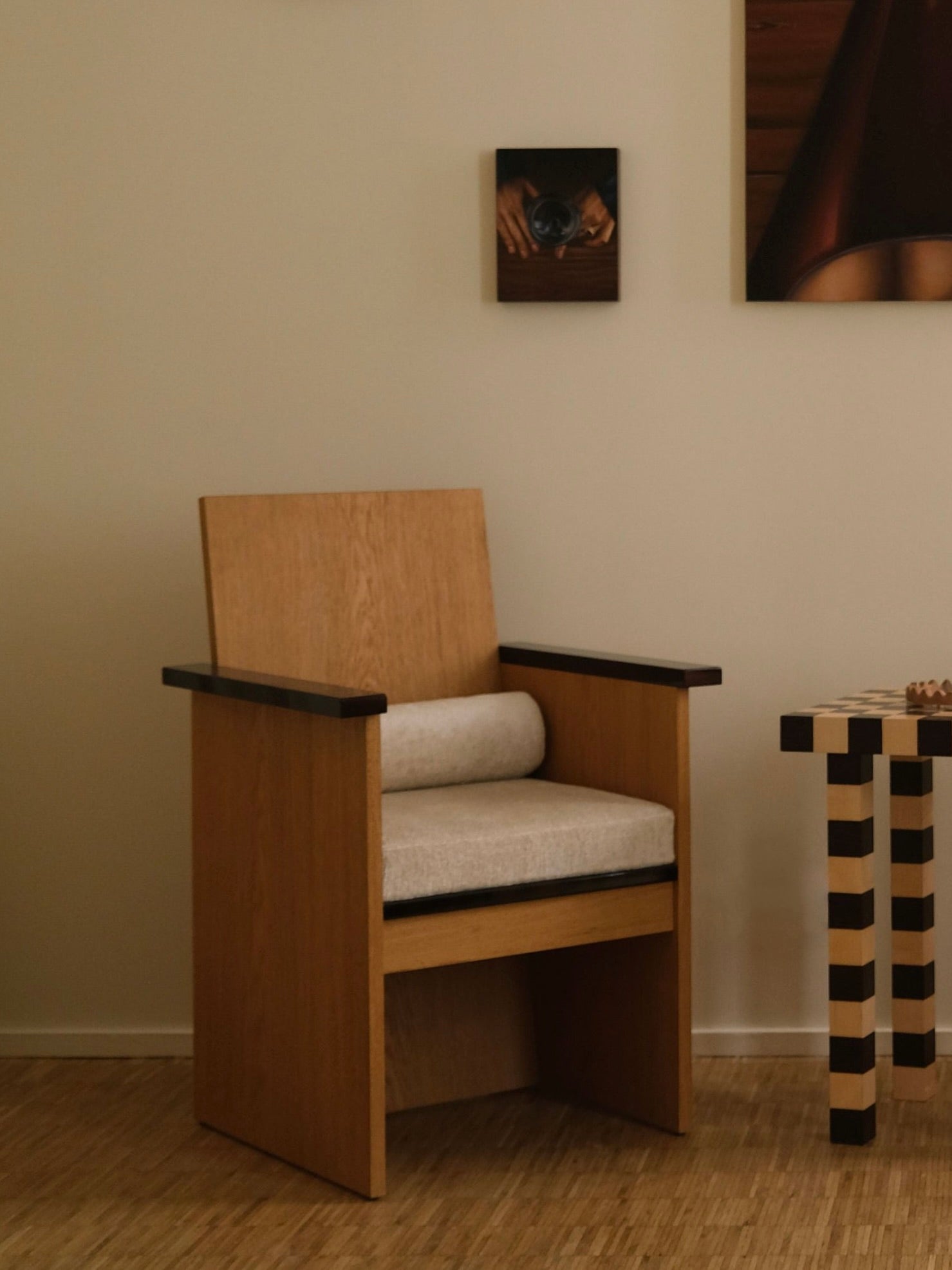 A minimalist room features the "Le Parfait" armchair by HAUVETTE & MADANI, with lacquered armrests and a cushion, a small checkered side table, and two artworks on the wall. The floor showcases a brushed oak texture, and the overall color scheme is neutral.