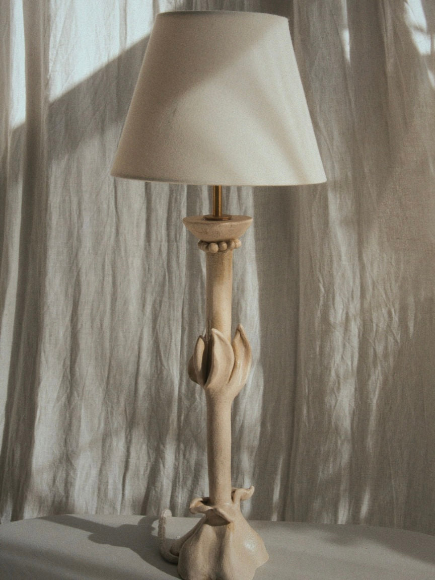 The "Bloom" Table Lamp by Lorea Palacios is a handmade piece crafted from chamotte white clay, featuring an abstract sculptural base with organic shapes. It stands in front of a textured light-colored curtain, topped with a simple white lampshade, and rests on a smooth surface.