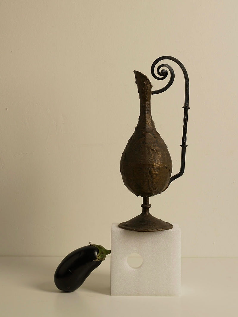 A tall, Swirl Iron Vase from Les Objets Amsterdam with a twisted handle stands on a white foam block with a round hole in it, exuding timeless elegance. A purple eggplant lies beside the block on a light-colored surface against a neutral background. The vase has a weathered, rustic appearance.
