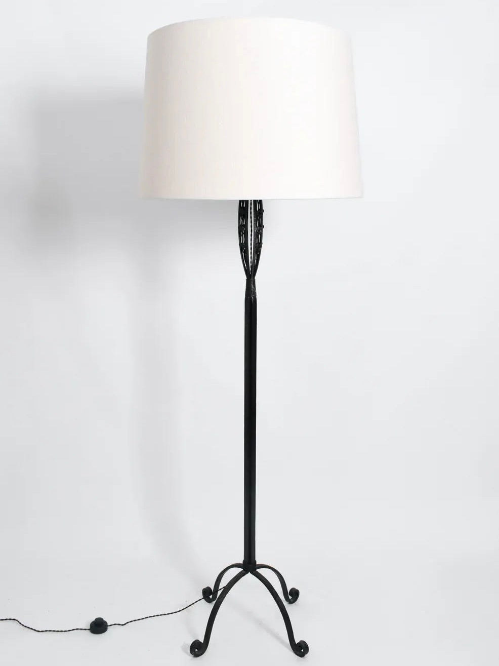 The Arts Deco Iron Floor Lamp from Barracuda Interiors adds a classic Art Deco flair with its tall black metal stand and white lampshade. Its base features elegant curved feet, while the electric cord extends from it with a round switch for convenience. Set against a plain light gray background, this sophisticated piece perfectly showcases Gingko decor style.