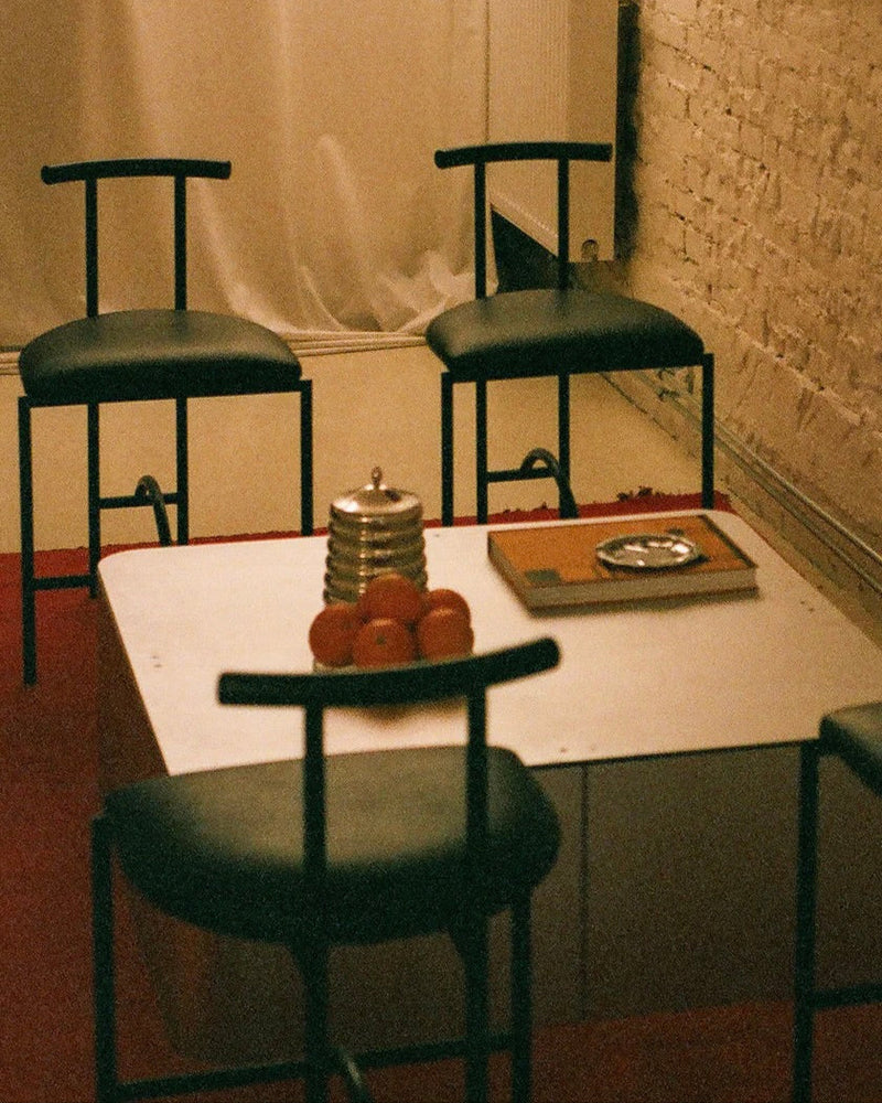 A minimalist room features four black chairs around the PARCTA 'Gadroon' Aluminium Coffee Table. On the table sit a stack of oranges, a round silver container, and a closed book. White brick walls and sheer curtains complete the serene setting.