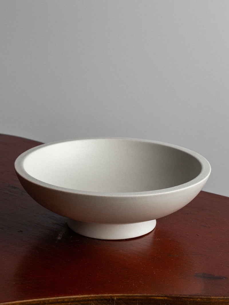 A simple, white 'Carrara' ceramic bowl from the 1940s Sweden collection by Spigel, featuring a wide rim and a sturdy base, sits on a wooden surface against a plain, light gray background. The minimalistic design of the bowl is influenced by Wilhelm Kåge's designs for Gustavsberg. The wood has a rich, warm hue, providing contrast to the clean lines of the bowl.