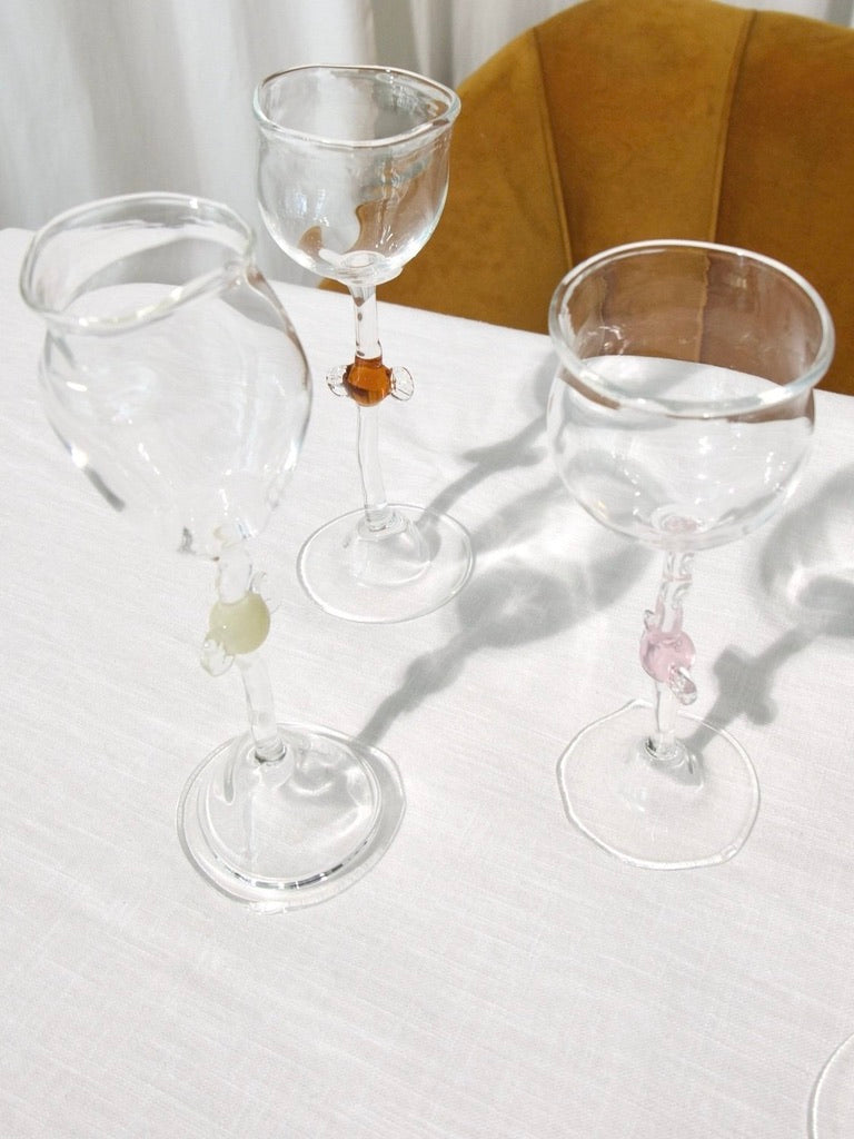 Three Vivian Alarcón Mouth Blown 'Candy' Wine Glasses with unique colorful stems sit on a white tablecloth by a mustard chair, their vintage-inspired shadows cast across the surface.