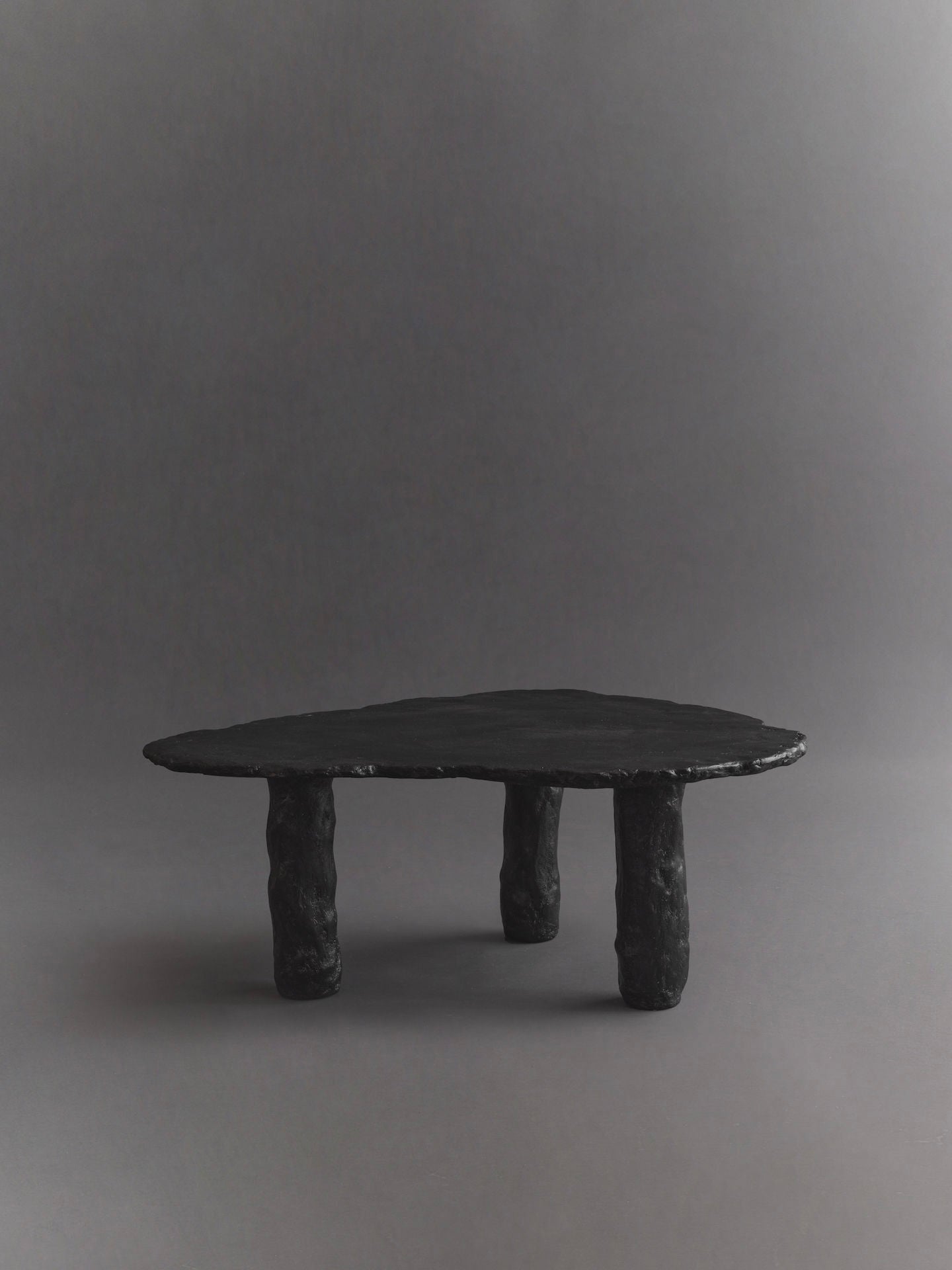 The Roca Nested Coffee Tables by Ombia Studio feature a unique, sculptural design with an irregular matte black top and three textured cylindrical legs, set against a neutral gray backdrop. This handmade piece adds modern elegance to any space.