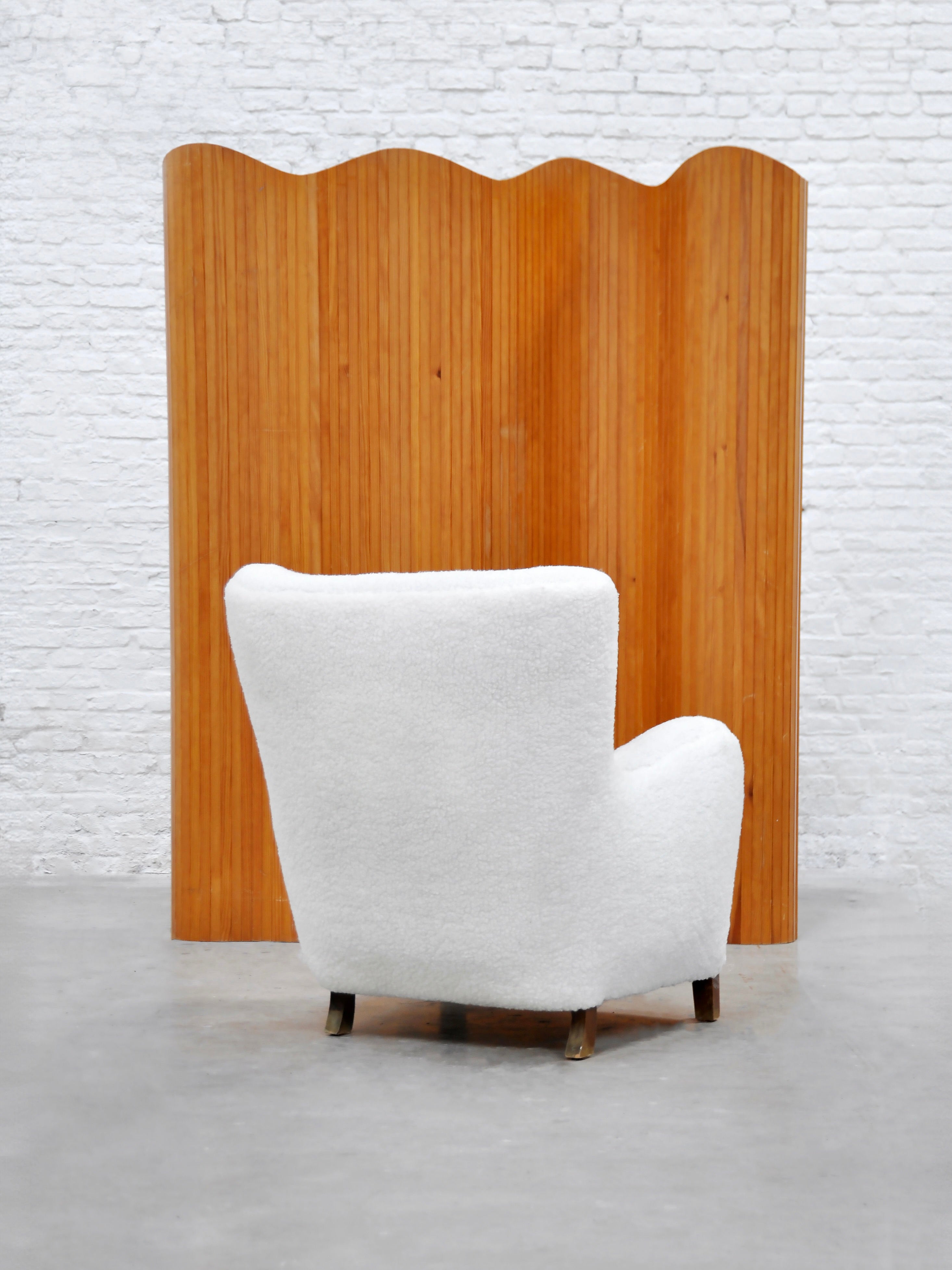 A white, plush armchair sits before the Jomaine Baumann Room Divider by Introverso, showcasing a tambour design. The minimalist setting boasts a light gray floor and white brick wall, echoing French Art Deco influences.