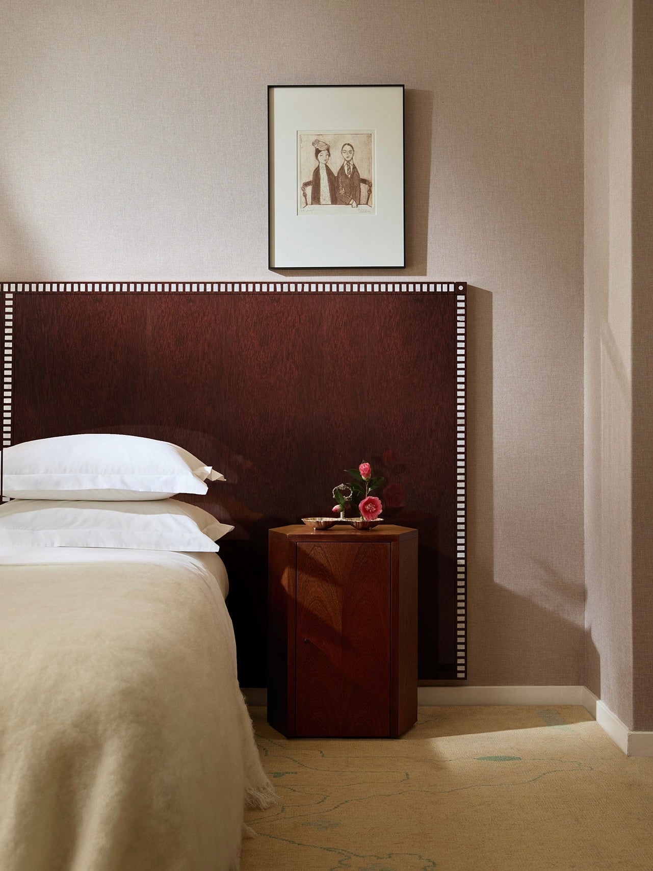 A cozy bedroom features a bed with white pillows and a beige blanket. The Aquarama headboard in African Mahogany by Lemon showcases a dark wood finish with a detailed border. Beside the bed, a wooden nightstand holds a small vase with flowers. Above the headboard is a framed black-and-white artwork.