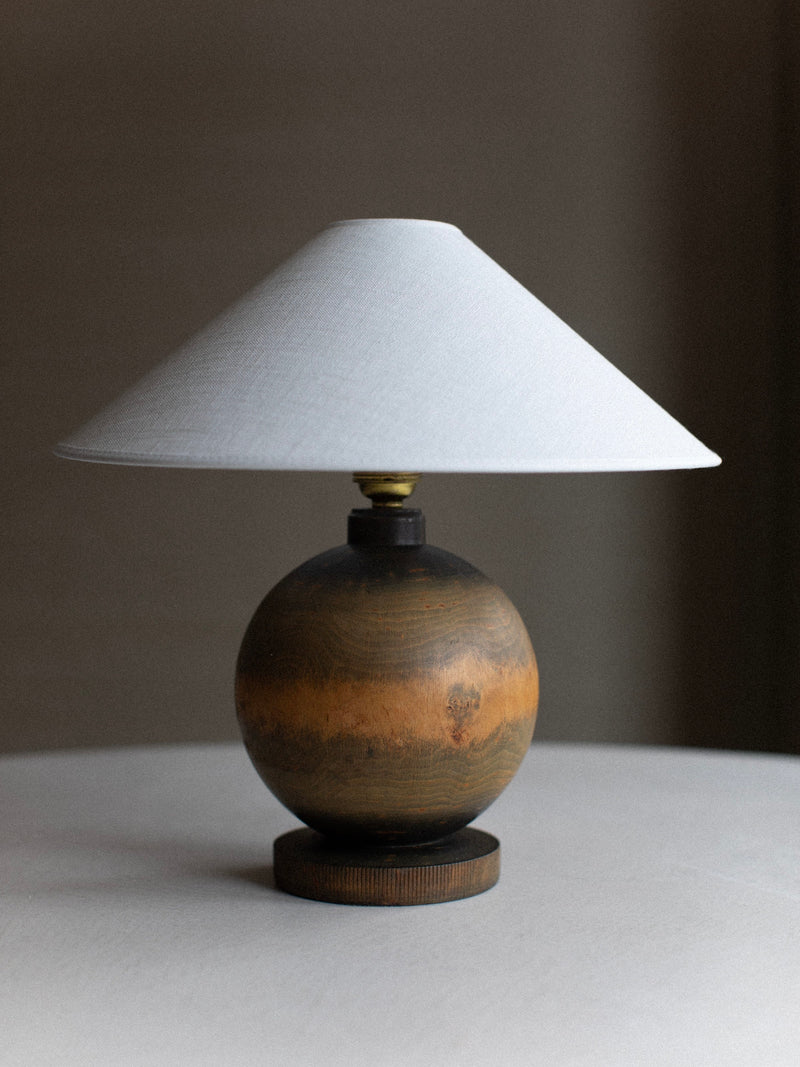 The 1920's Globe Lamp by Out For Lunch, featuring a round, rustic wooden base and a conical, white lampshade, is placed on a white surface. The worn, distressed wood finish adds to its vintage decor aesthetic. The background is a neutral, shadowy grey.
