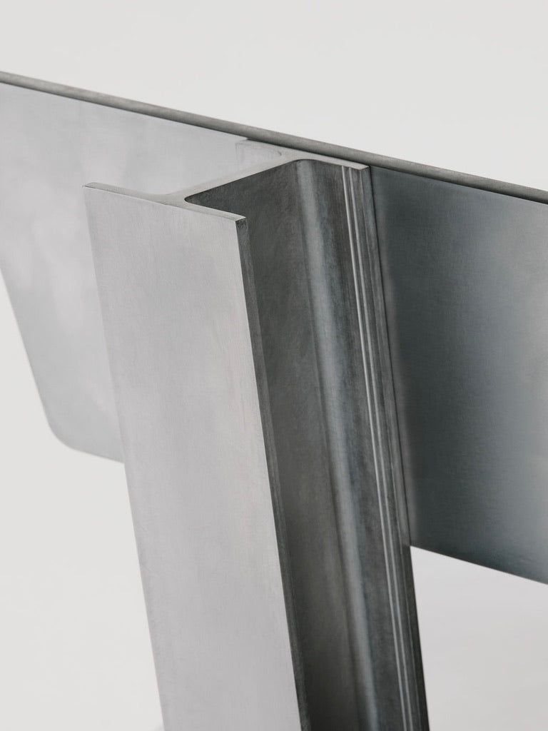 Close-up of Marquel Williams' Beam Chair highlights a modern metal structure with intersecting rectangular and angular elements. The brushed texture mimics I-beams, presenting a minimalist design against a white background.