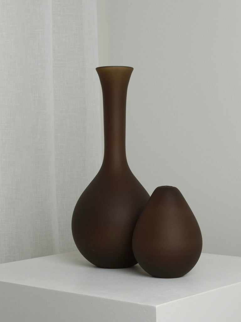 Two brown ceramic vases from Verre d'Onge, one of which is the XLarge Vase in Chocolate, are placed on a white surface. The taller vase with a narrow neck is mouth blown, while the shorter and rounder one boasts a rich chocolate hue. The background remains minimalistic with soft white curtains, contributing to the clean and simple aesthetic of the image.