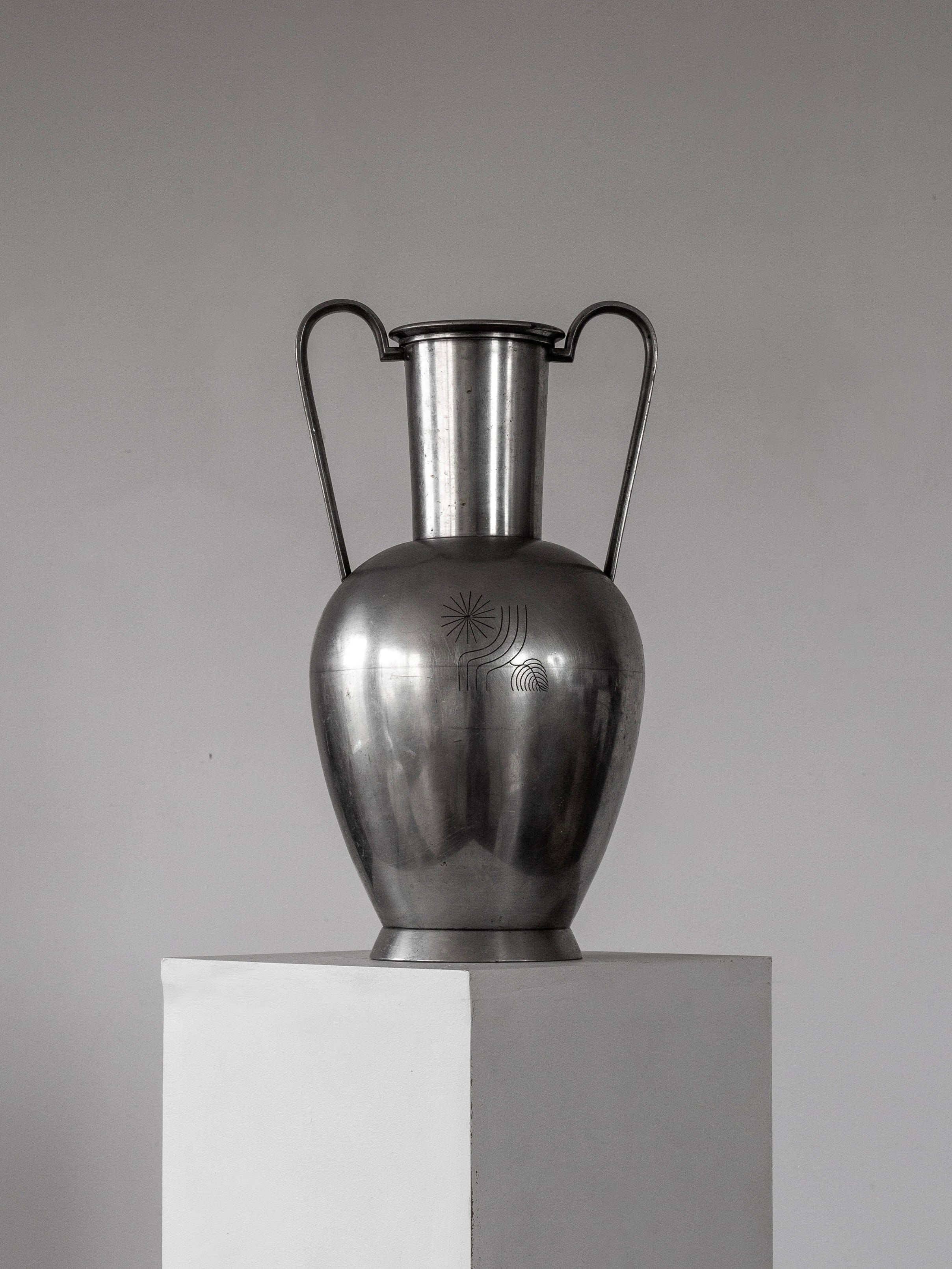 A Spigel large modernist metal amphora from the 1930s, reflecting Scandinavian style, stands on a white pedestal. It has two arched handles and subtle engravings, possibly influenced by Rolf Engströmer, against a plain gray background.