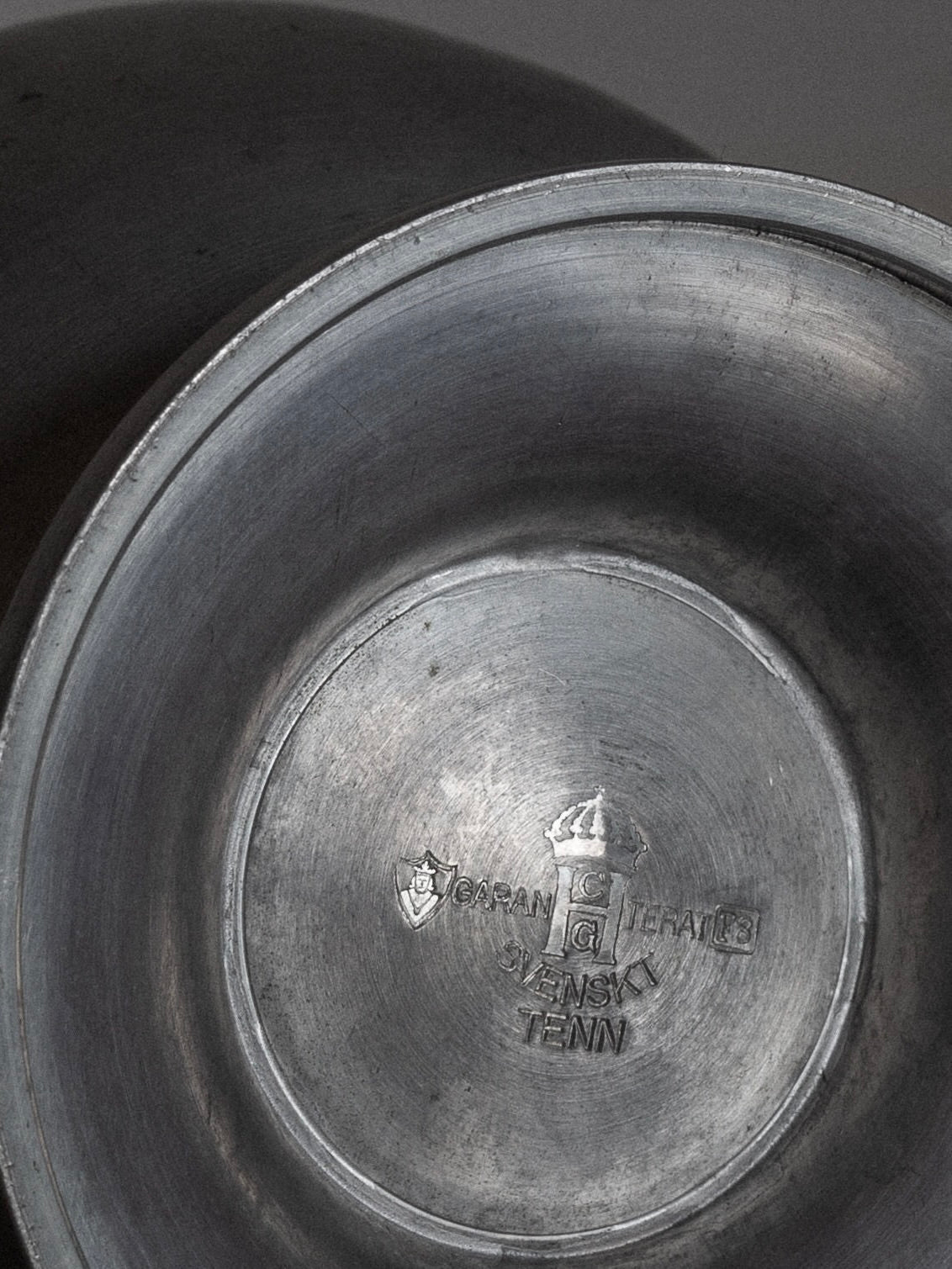 A close-up shot of a collectible Art Deco metal vase from the 1930s by Spigel, displaying engravings at its base. The engravings include a shield, a letter "G," a crown, and text reading "TSAGA T GRALTB," "SWENSKY," and "TENN." The slightly worn finish of the bowl exhibits elements of Swedish Art Deco style.