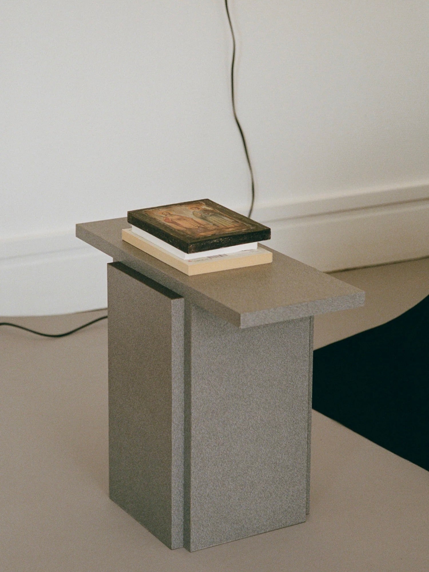 A "Mara T" Side Table – Silver Noise, crafted by TEGET and made of durable materials, is positioned against a plain wall. Atop the small side table rests a book and a framed picture, with a black cord noticeable in the background.