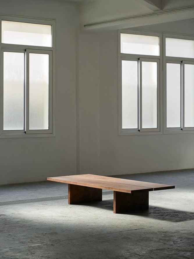 A minimalist room with large frosted windows allows soft natural light to shine upon the ASERIES OF OBSTACLES, SL Tanizaki Coffee Table. The simple table, reminiscent of Japanese living rooms, casts a shadow on the concrete floor, serving as the serene space's central focus.