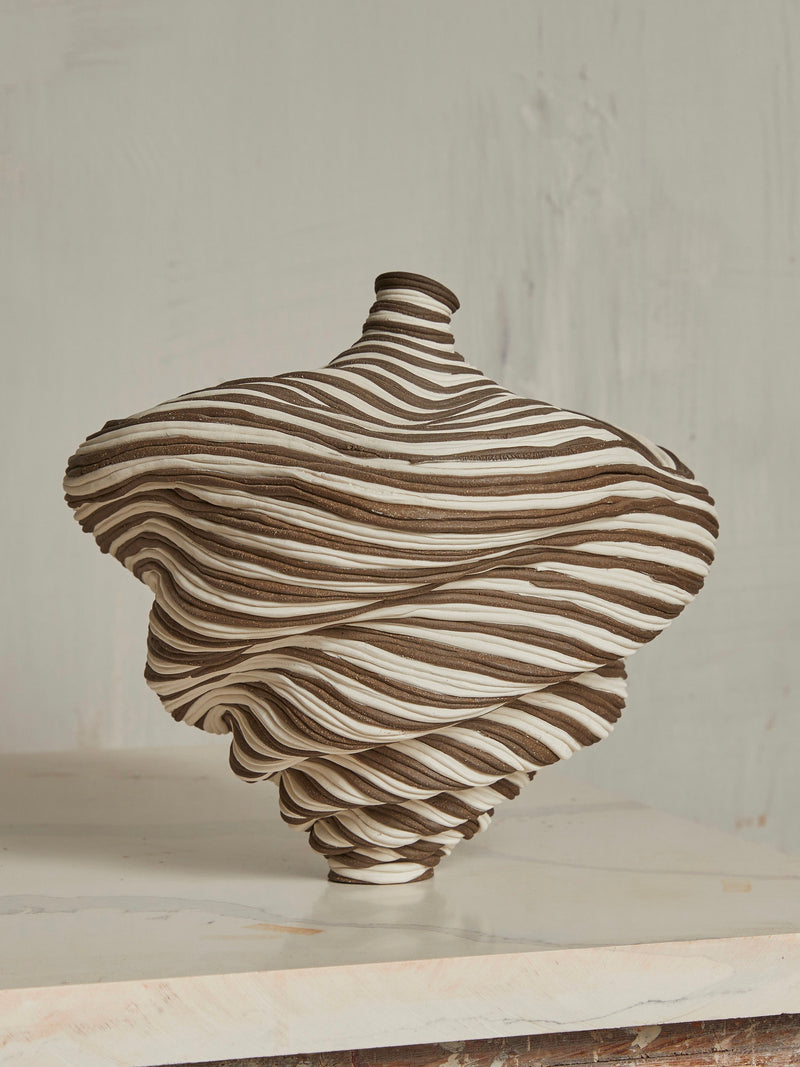 The Soucy Shop's Black & White Bottle is a unique sculptural ceramic vase with swirling brown and white stripes. Crafted from stoneware coils, its dynamic shape resembles a spinning top, displayed on a light surface against a muted backdrop.