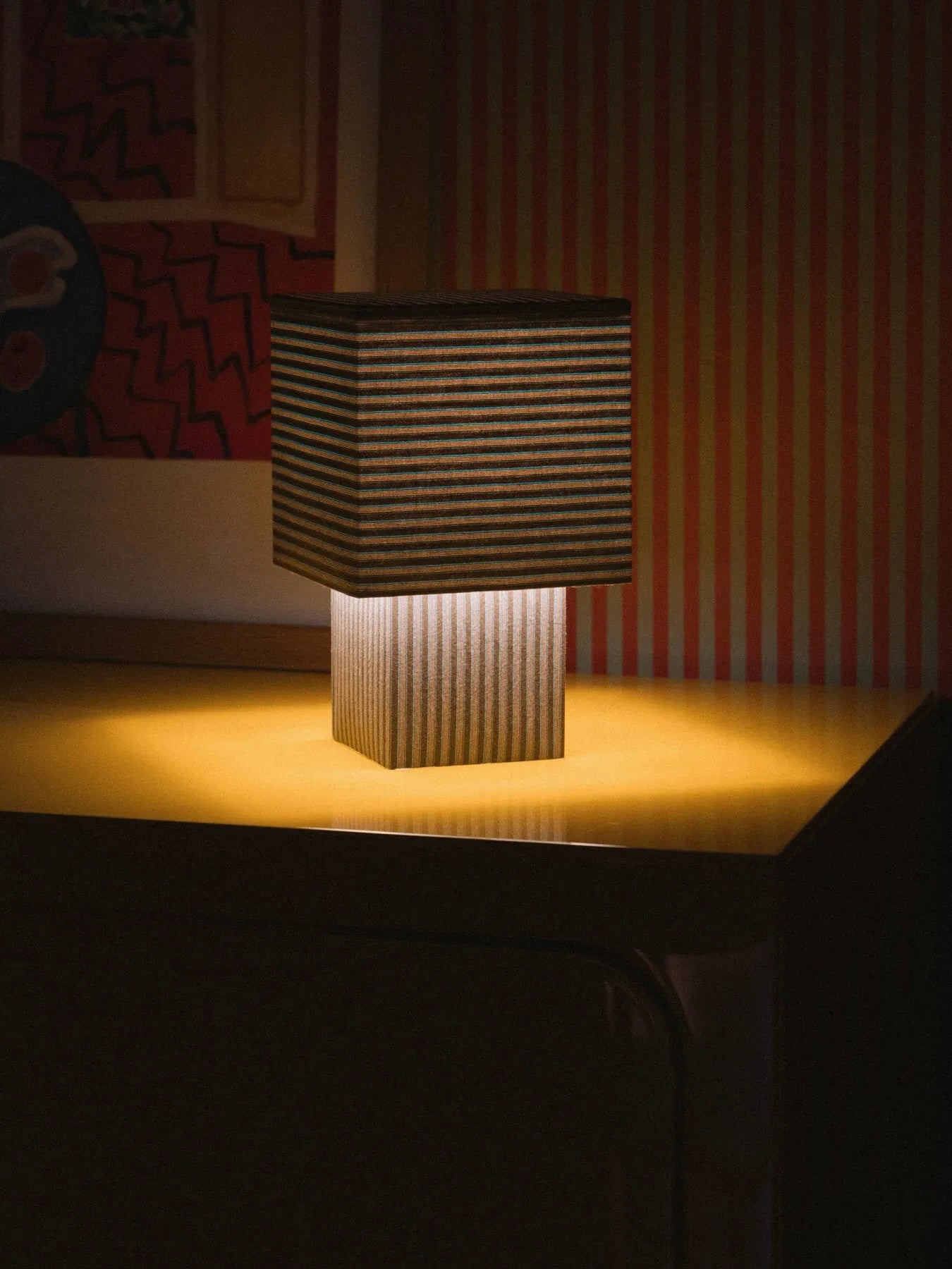 The "La Petite Boîte" table lamp by mattina moderna features a rectangular, striped shade crafted from limited edition deadstock fabrics. It emits a warm glow that beautifully illuminates a room adorned with patterned wallpaper and framed art. The handcrafted lamp rests on a reflective golden-yellow surface, enhancing the cozy ambiance.