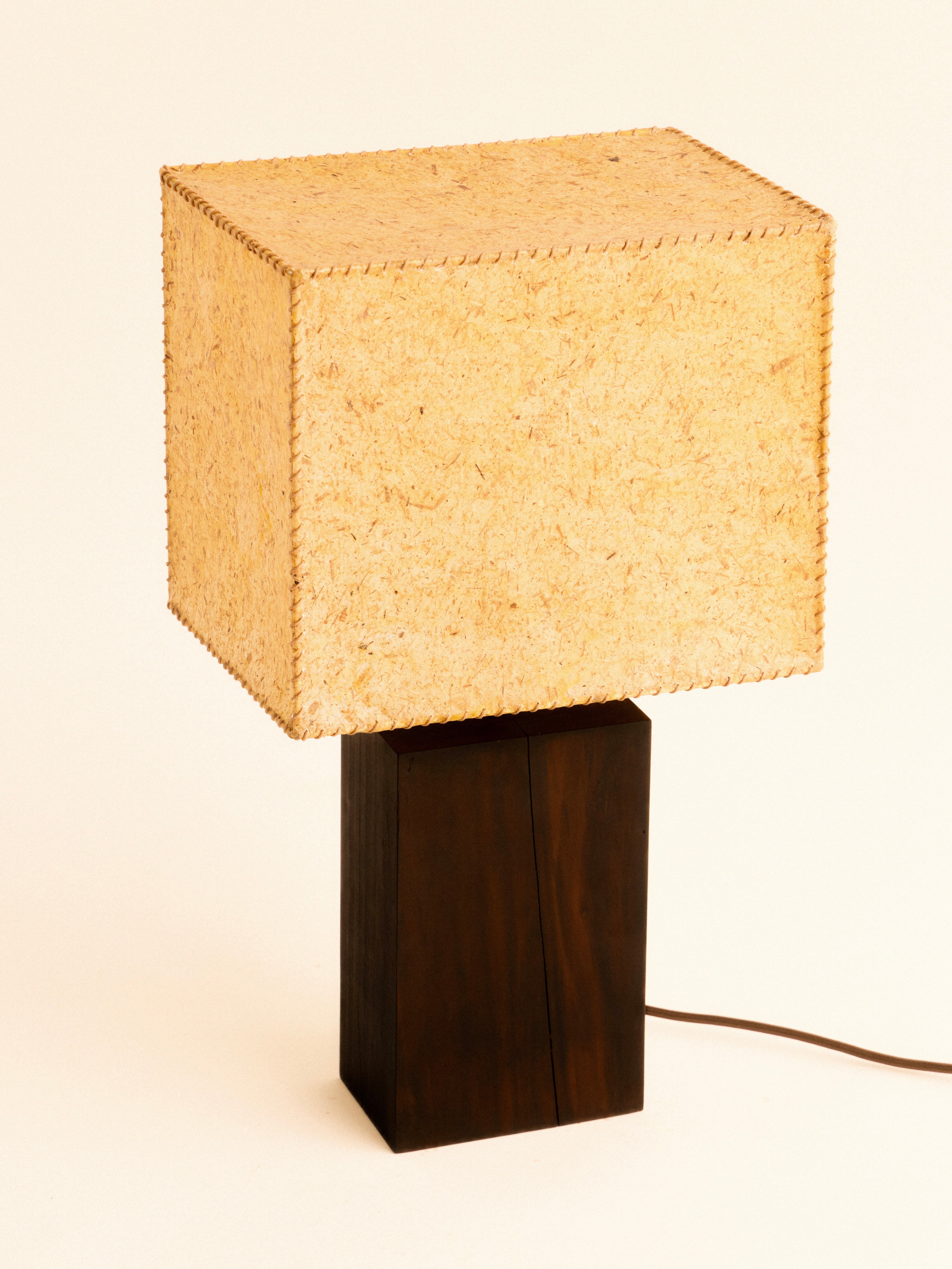 The 1948 Table Lamp by Lana Launay showcases a handcrafted square design complemented by a beeswax shade, set on a rectangular base made of dark wood. The power cord extends from the rear, all placed against a simple off-white background.