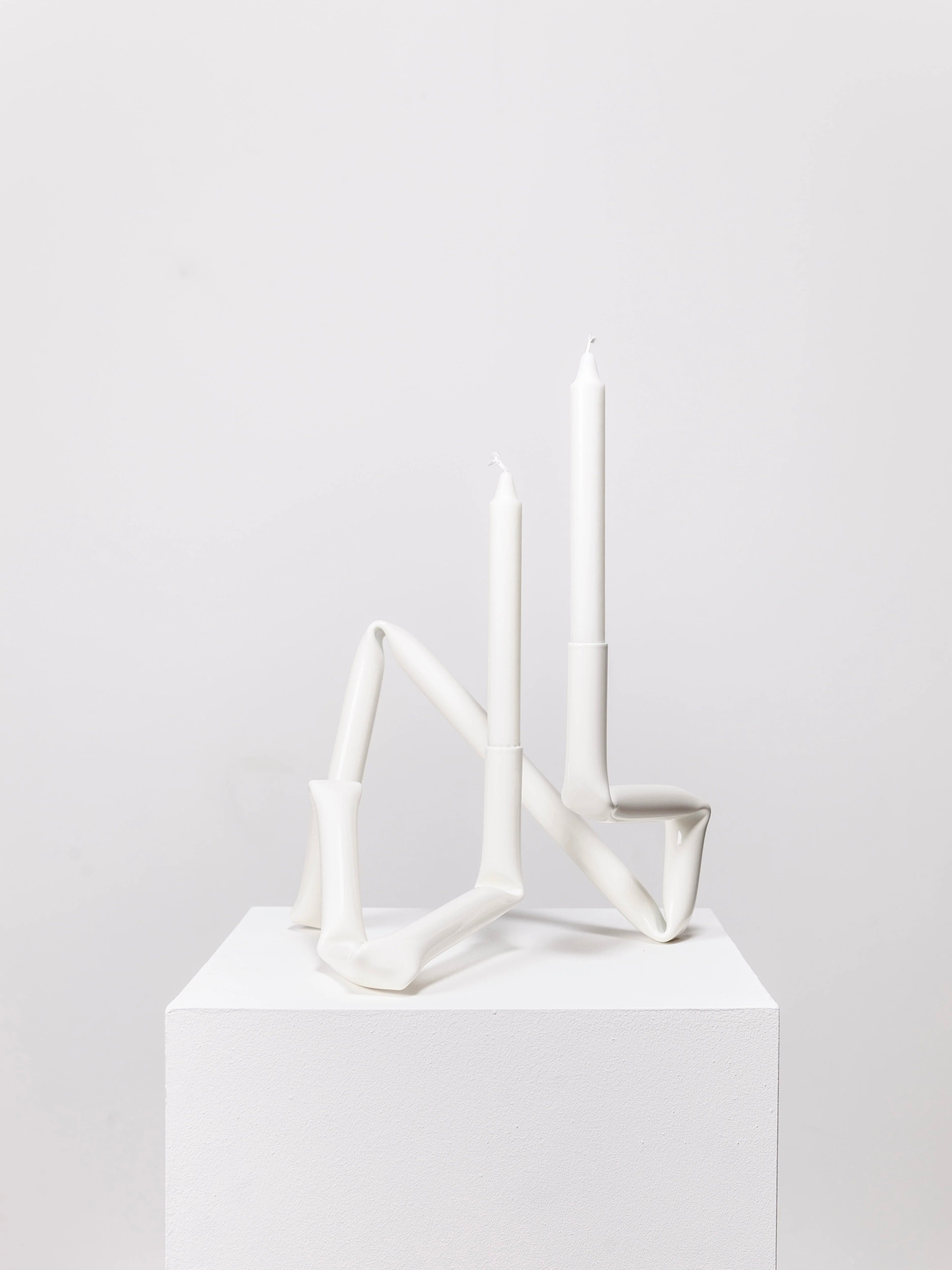 Two white, asymmetrical, hand-crafted Bucatini candle holders from AOT STUDIO are displayed on a white pedestal against a plain background. Each holder supports a tall, white taper candle, one slightly shorter than the other. These exquisite pieces are part of the Primi collection.