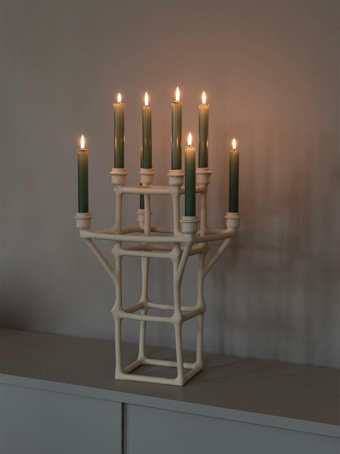 The Clay Chandelier by Solenne Belloir is a modern piece with an abstract, geometric design that holds eight lit green candles, casting a warm glow. This handcrafted chandelier sits on a simple grey surface against a soft grey wall, providing a truly unique ambiance reminiscent of minimalist elegance.