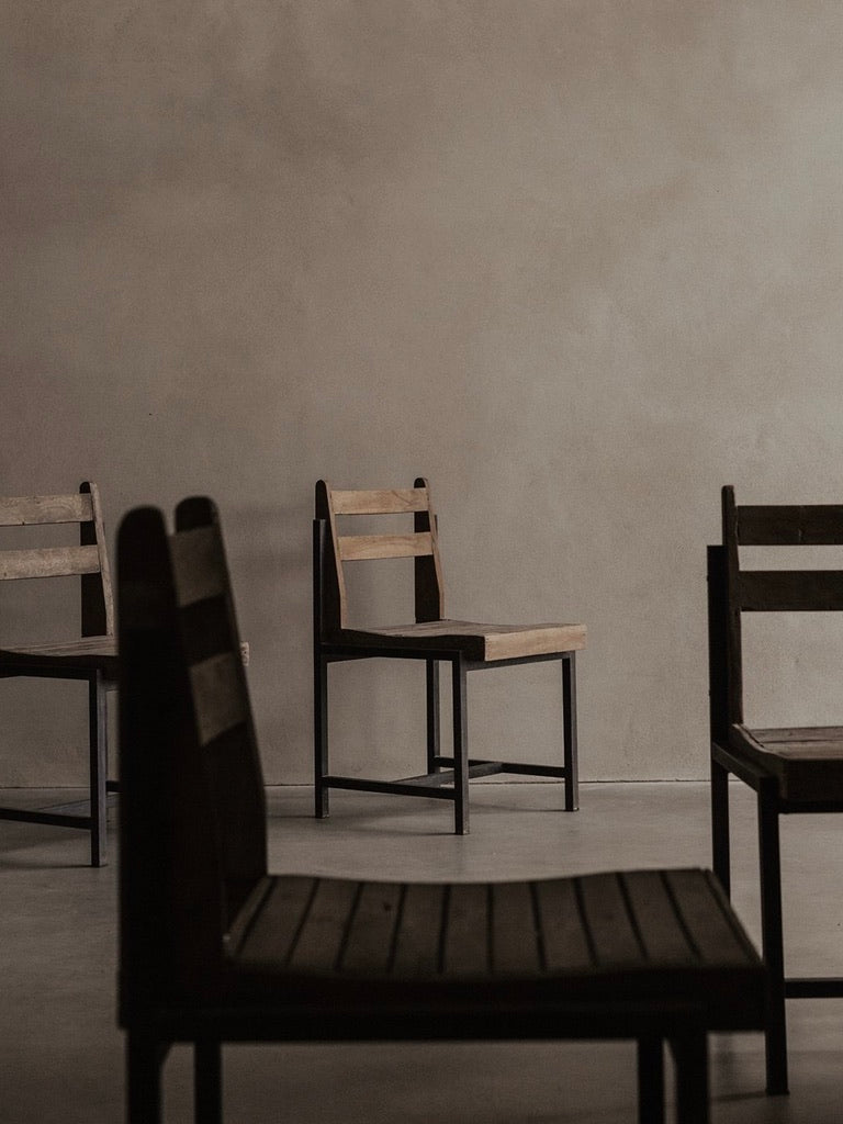 In a minimalist room, four Bicci de' Medici Pierre Jeanneret Authentic PJ-SI-65-A chairs adorn the concrete floor. The plain, textured walls offer a serene backdrop, while soft lighting highlights the simplicity of teak wood slats, enhancing tranquility.