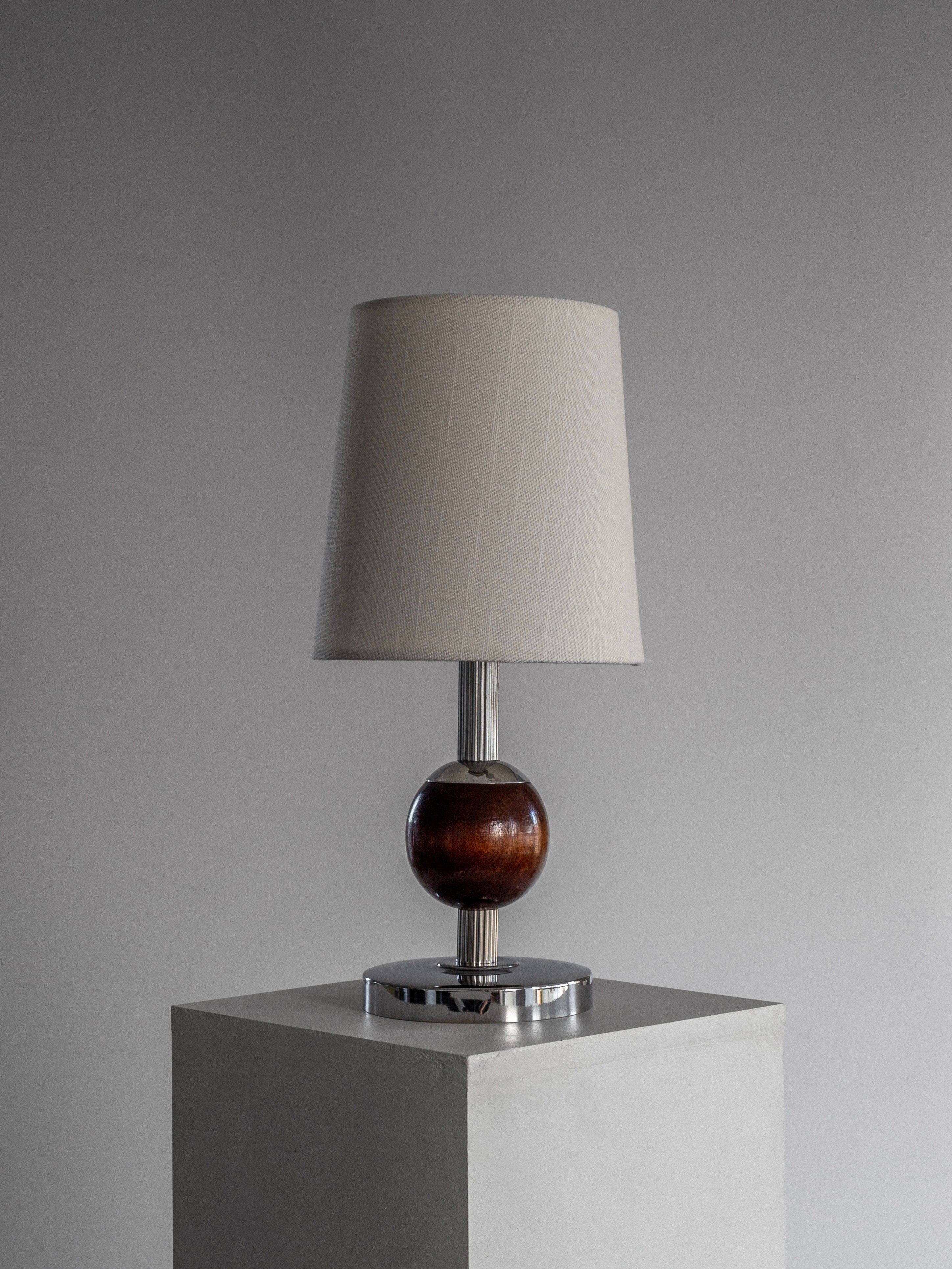 The Spigel Wood & Chrome Table Lamp from the 1930s gracefully sits on a chrome and wood base, featuring an elegant beige shade. This Art Deco piece captures the essence of 1930s Sweden, perfectly poised on a gray pedestal against a neutral gray backdrop.