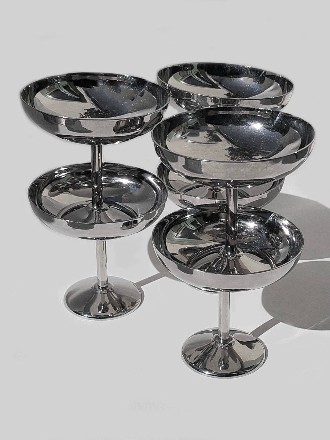 A set of six Les Objoies Ice Cream Cups, made from stainless steel with reflective surfaces, are arranged against a plain white background. Each cup features a round bowl atop a slender pedestal, perfect for serving ice cream. The cups vary slightly in height, creating a tiered effect.