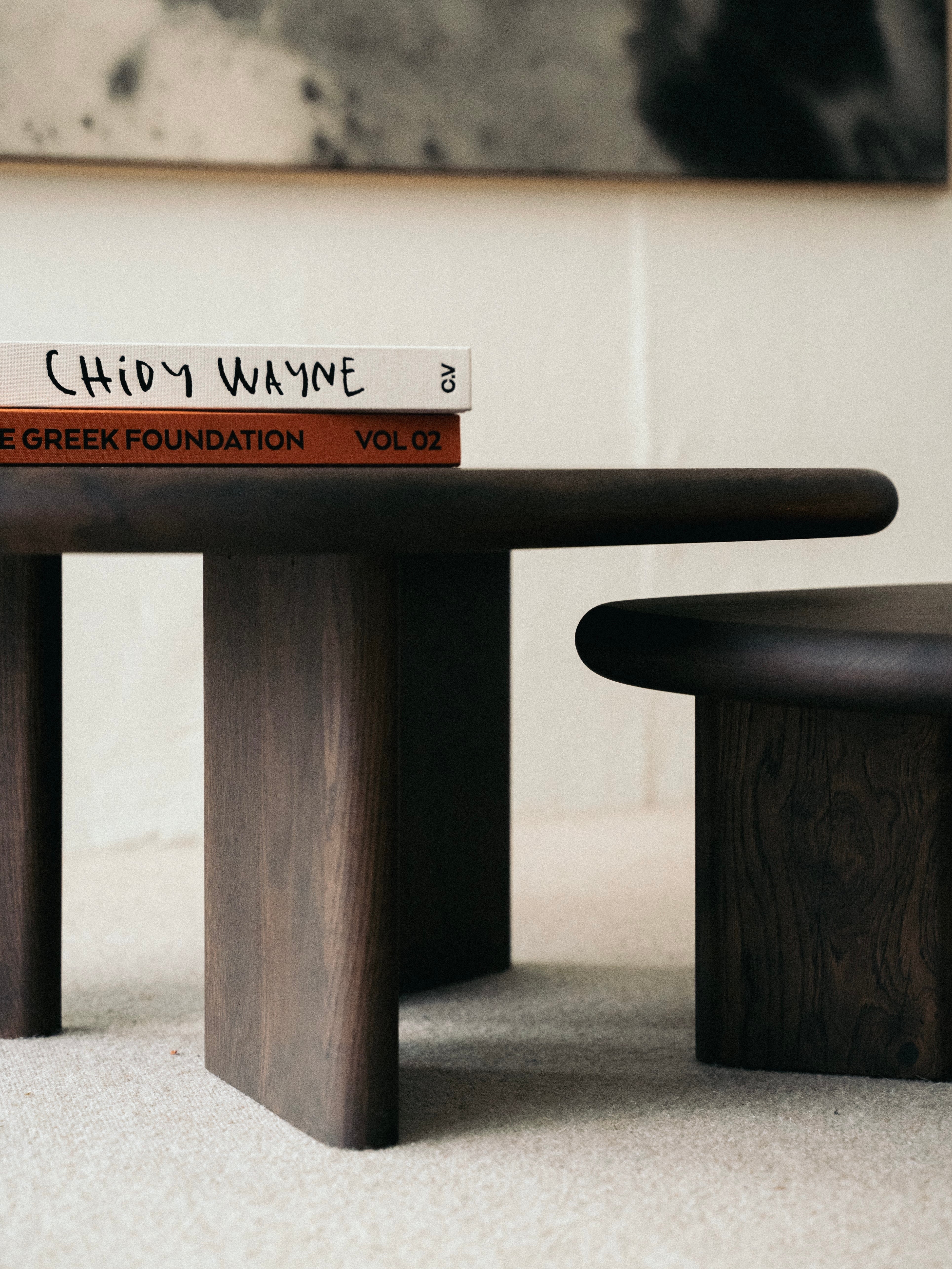 A close-up of the KØGE Design Double Espresso Coffee Table, adorned with an espresso-tinted varnish finish. The table features two stacked books with partially visible covers; one book titled "Chidy Wayne," and the other "The Greek Foundation, Vol 02." The setting exudes a minimalistic and modern aesthetic.