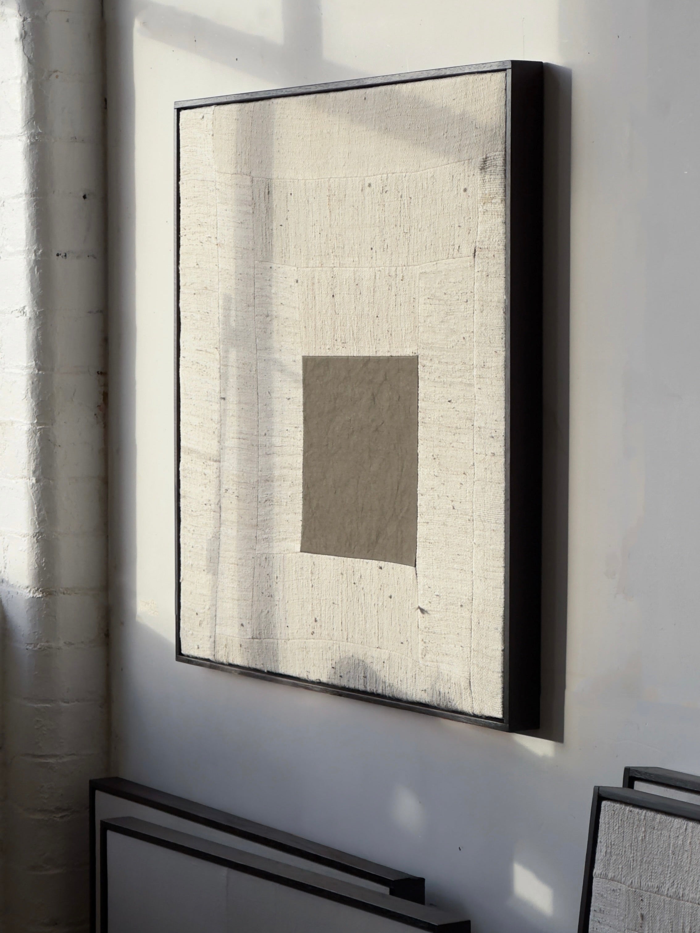 Are You Open?", 2025, by Bec Kirby features a minimalist masterpiece: a large light beige square of handwoven kapok fibre cloth with a smaller gray square at its center, framed by solid oak. Sunlight casts gentle shadows, highlighting the layered textures and captivating elegance of this artwork.