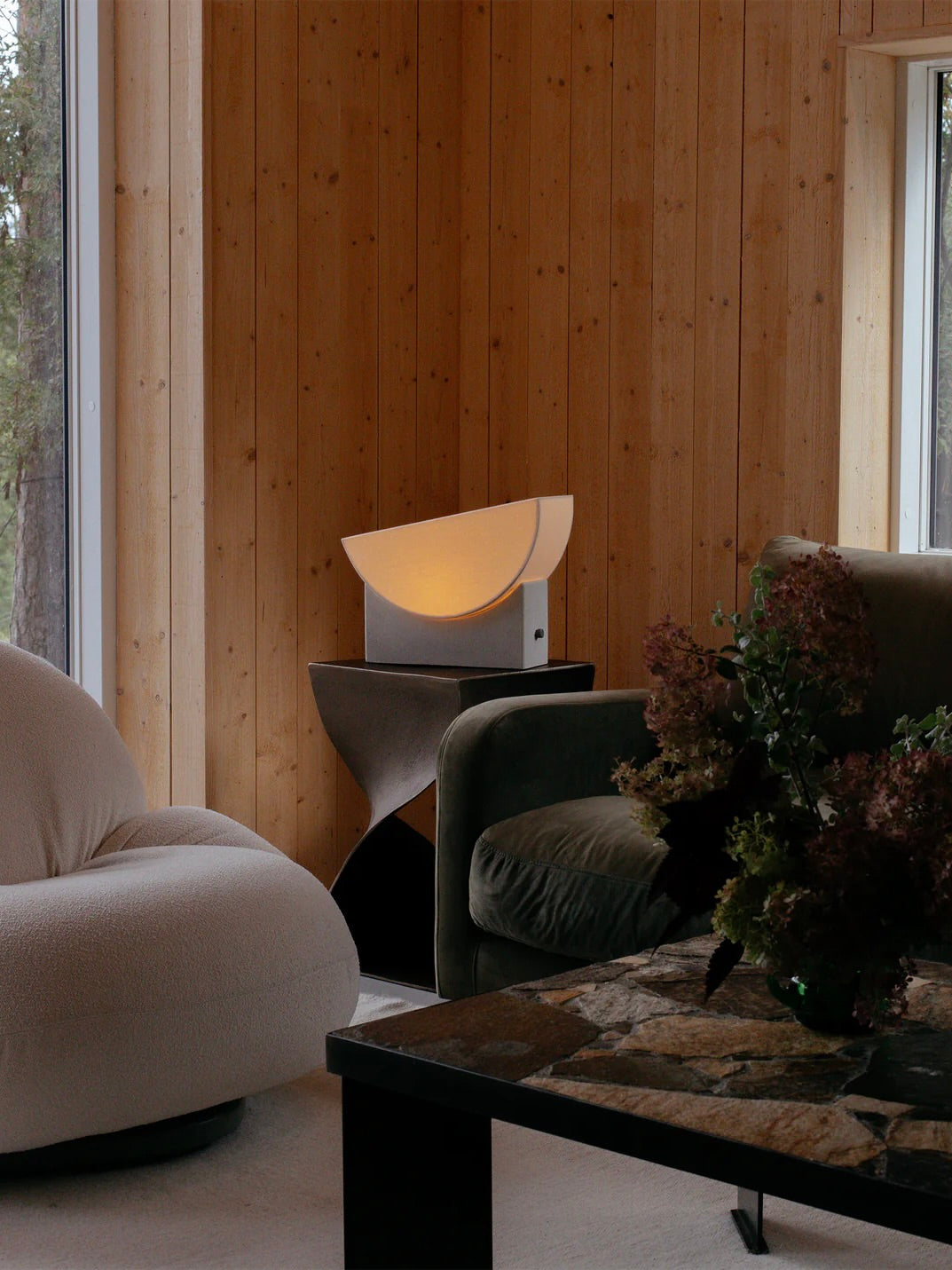 The warm living room, adorned with wooden walls, showcases the "Reminder" Table Lamp by Enkei Lights AB on a geometric side table. The space also includes plush white and green sofas crafted from deadstock textiles, complemented by a stone-top coffee table, while a window offers a view of lush greenery outside.