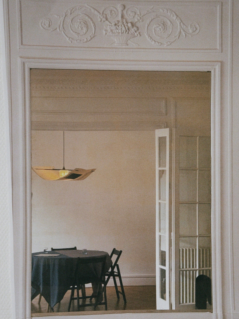 A mirror reflects a dining room featuring a handmade Sail Lamp – Rice Field by TEGET above a round, dark-clothed table. The room includes ornate molding, partially open French doors, minimalist decor, and unique paper collage art.