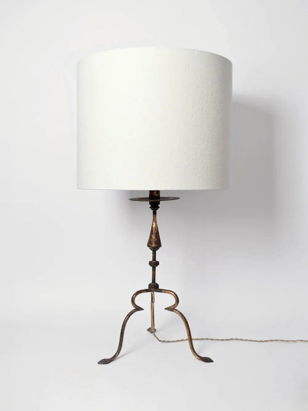 Introducing the Barracuda Interiors Golden Iron Table Lamp from the 1960s, this vintage piece features a large white cylindrical shade perched atop an intricately crafted gilded hammered iron stand with three elegantly curved legs. The lamp emanates a charming vibe reminiscent of 1960s Spain, complete with a cord extending from its base against a plain white backdrop.