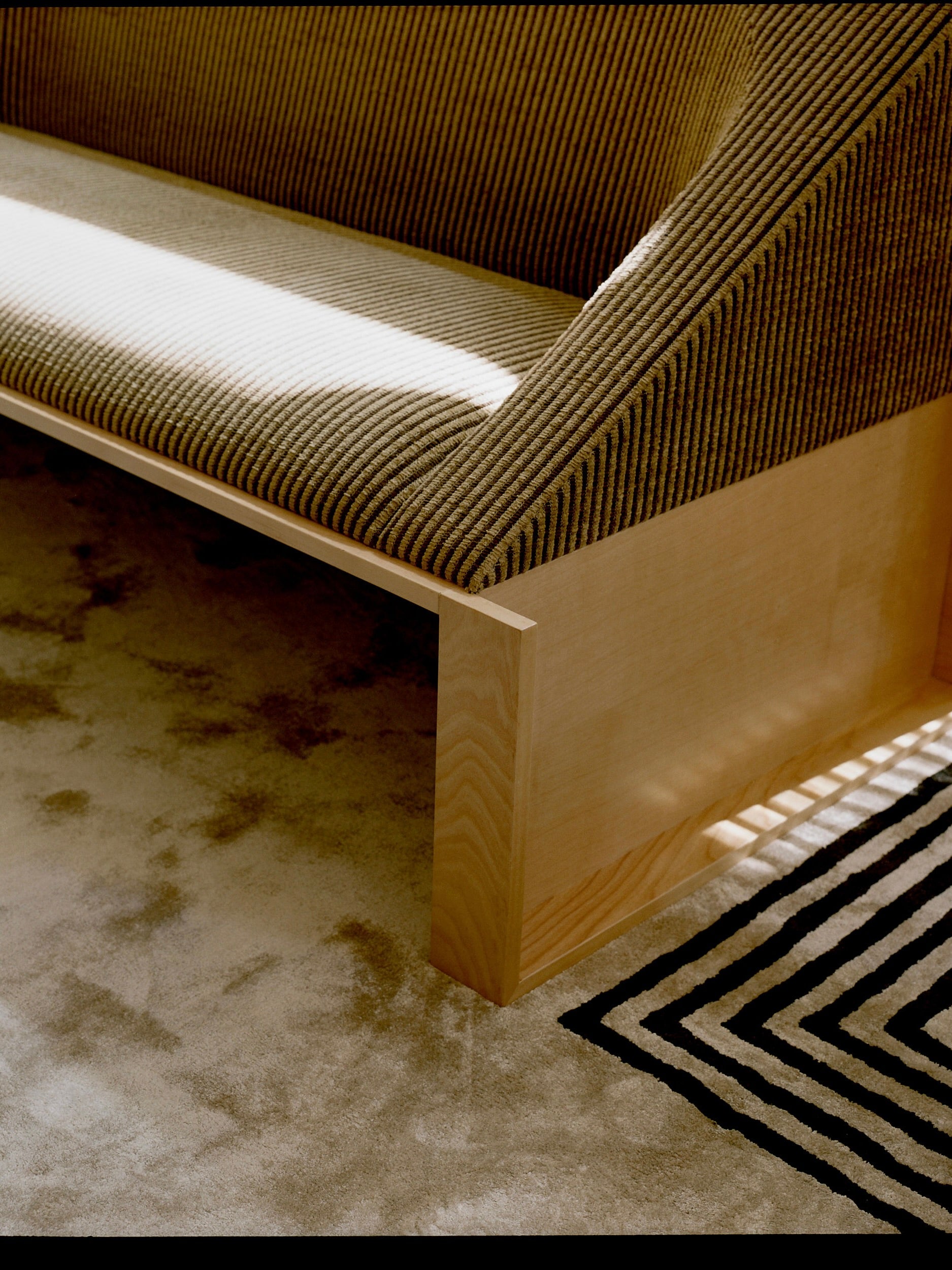 A close-up of the 'Perceptions' Sofa Bench by Known Work reveals its modern, minimalist wooden design with a textured, striped fabric seat. It sits on a light-colored carpet with a geometric black and white pattern, where sunlight creates dynamic shadows across the furniture and floor.