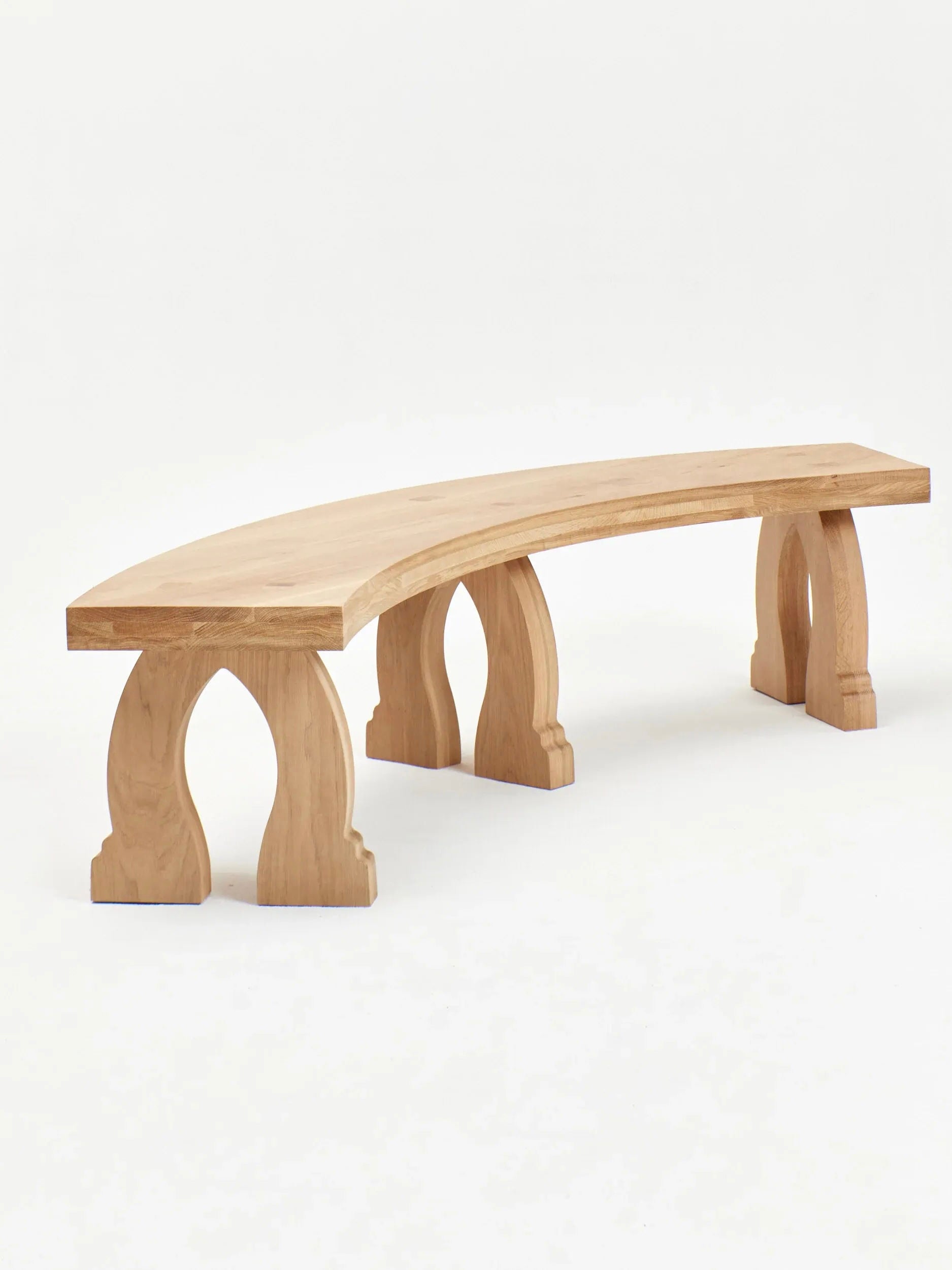 The "Sintra" Curved Bench in Oak by Project 213A is a stylish piece featuring a minimalist design with a gently curved seat and three intricately carved legs. Expert artisans have crafted the arch-like cutouts on the legs, adding an elegant touch to the natural wood finish against a plain white background.