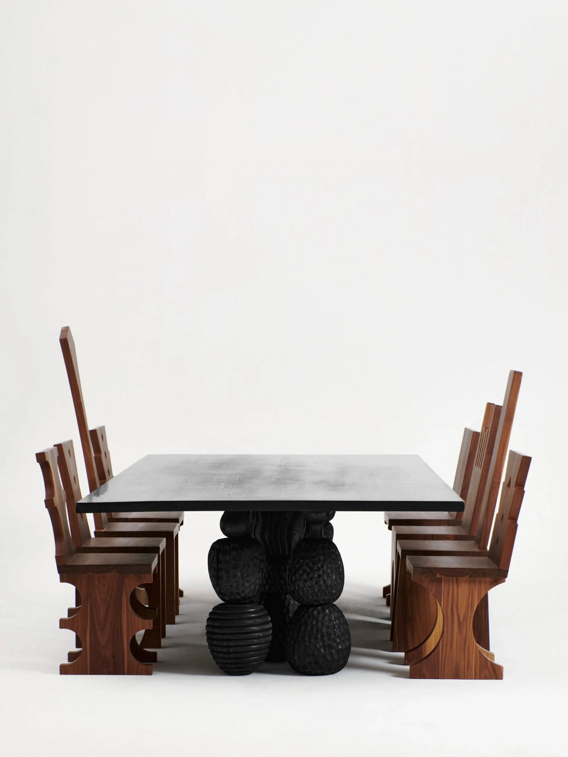 A sleek, rectangular Menina Dining Table by Project 213A with a black, sculptural base is complemented by six matching handcrafted chestnut wooden chairs. These chairs feature intricate geometric designs on the legs and backrests, all set against a plain white background.