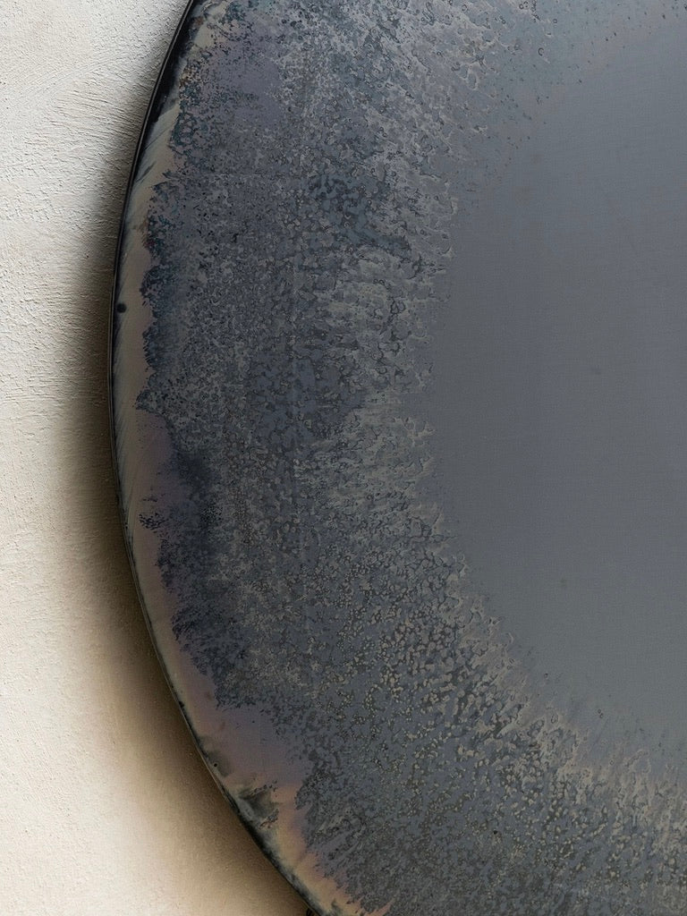 The HOMA Gray Mist Convex Oxidized Mirror features a round, textured metallic surface with a gradient of smoke gray to lighter shades. Its smooth edge contrasts beautifully with the rough textures within.