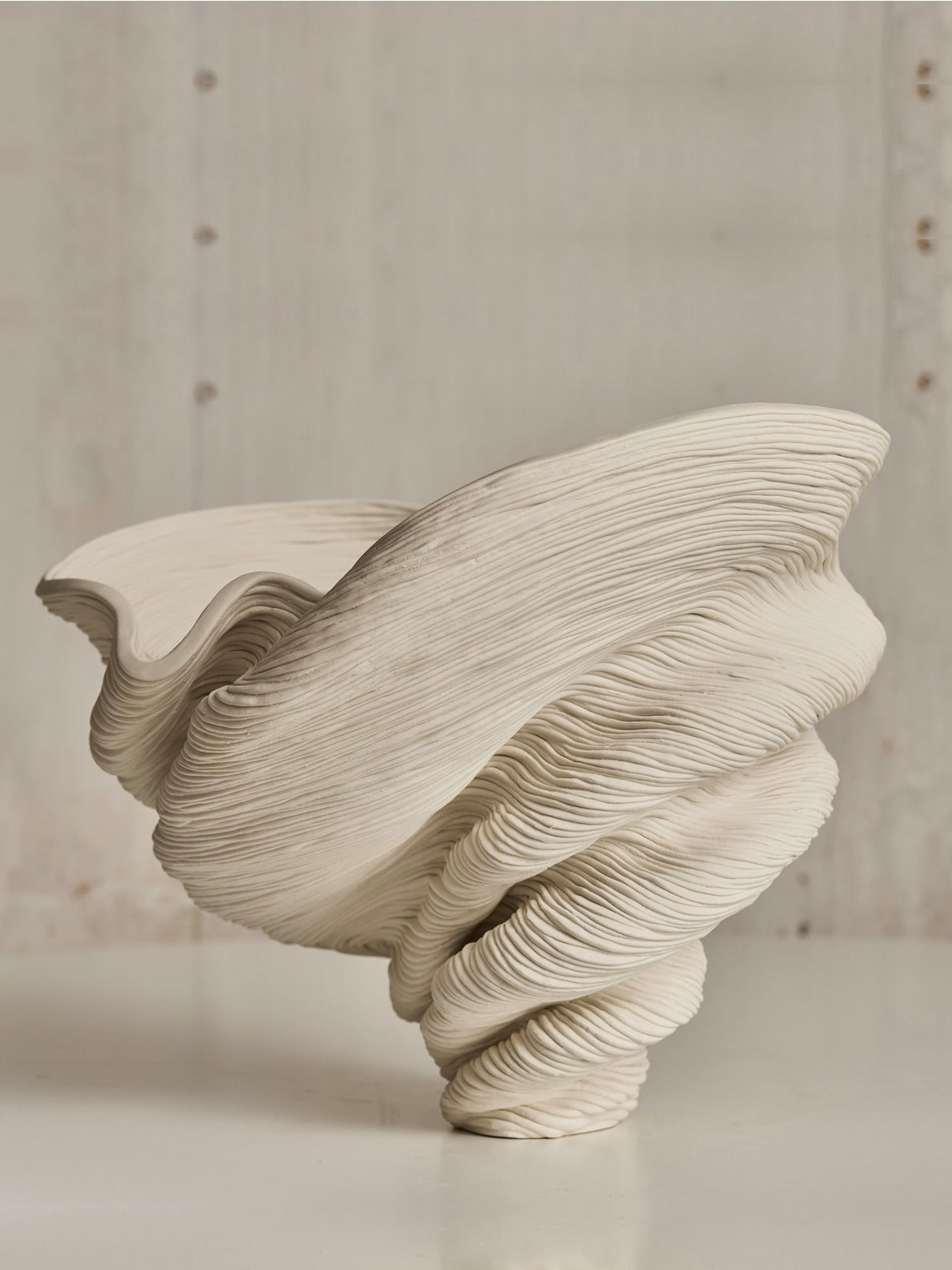 The Soucy Shop's Medium Flutter is a handmade sculptural piece crafted from layered porcelain coils, forming a fluid, swirling spiral reminiscent of a twisted shell. It's displayed on a plain surface with a neutral background.