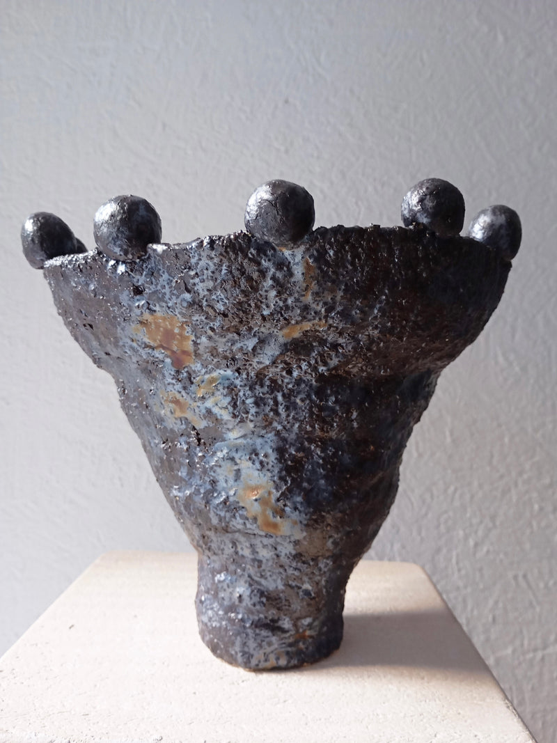 The "Coupe Lilibeth 2" by Claire Pain is a sculptural ceramic vase with a textured, metallic glaze. Its conical shape widens at the top and features rounded protrusions around the rim, ideal for decorative use against a subtle, textured background.